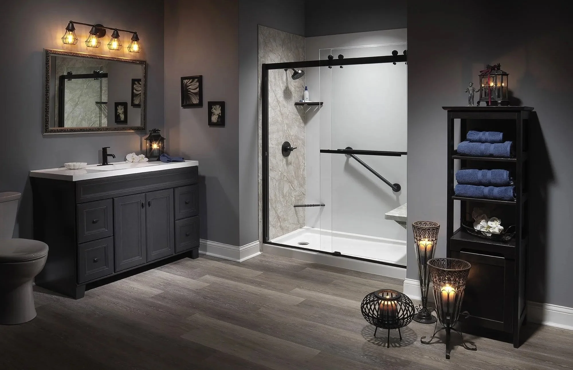 Modern bathroom with grey walls, a black vanity with a white countertop, a large mirror, framed wall art, a walk-in shower with glass door and marble accents, and black shelving with neatly folded blue towels. Decorative candles and lanterns are also
