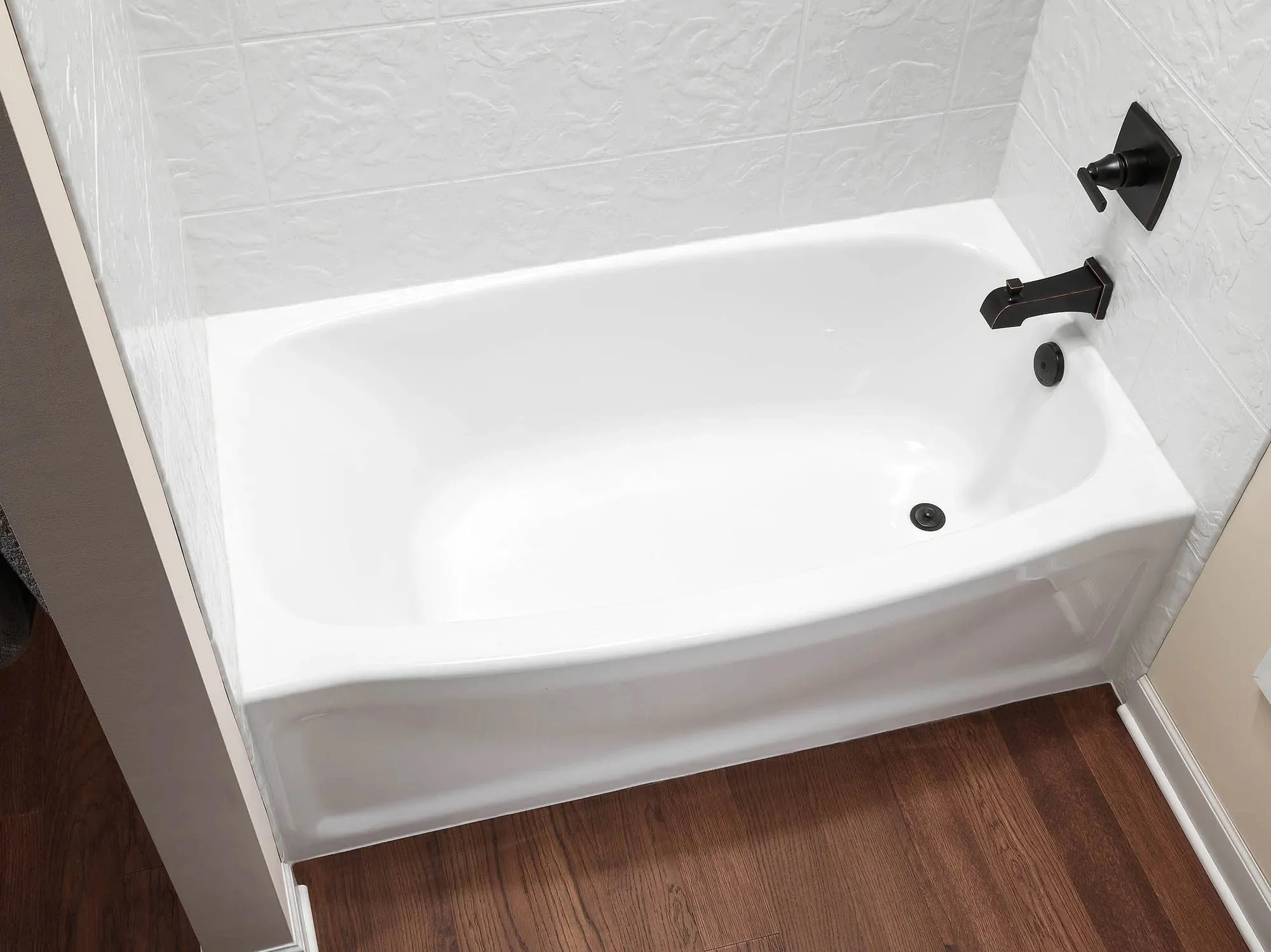 White bathtub with black fixtures and a bronze faucet in a bathroom with white textured wall tiles and hardwood flooring.