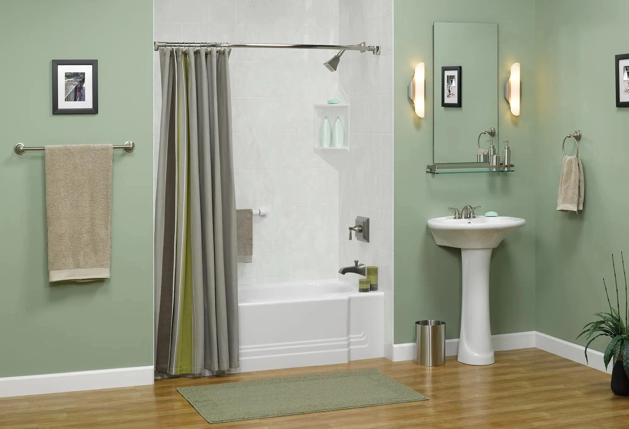 Bathroom with green walls, white bathtub with shower curtain, pedestal sink, framed pictures, beige towels, wall-mounted lights, mirror, small shelf, decorative plant, wooden floor, green rug, and small shelves with toiletries.