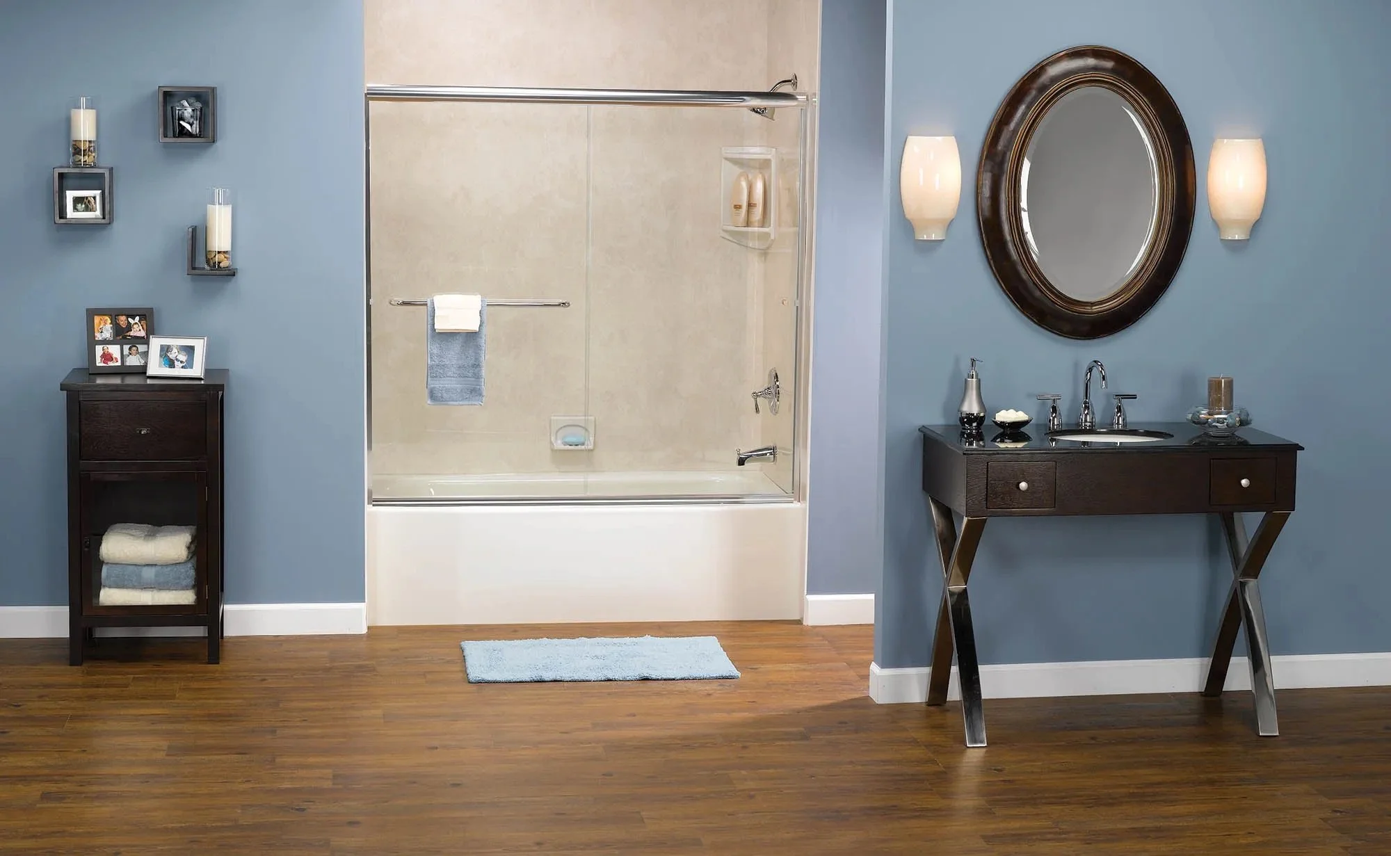 Modern bathroom with blue walls, a bathtub with glass sliding door, a dark wood vanity with a round mirror, wall-mounted light fixtures, a small dark side table with photos and towels, and hardwood flooring.