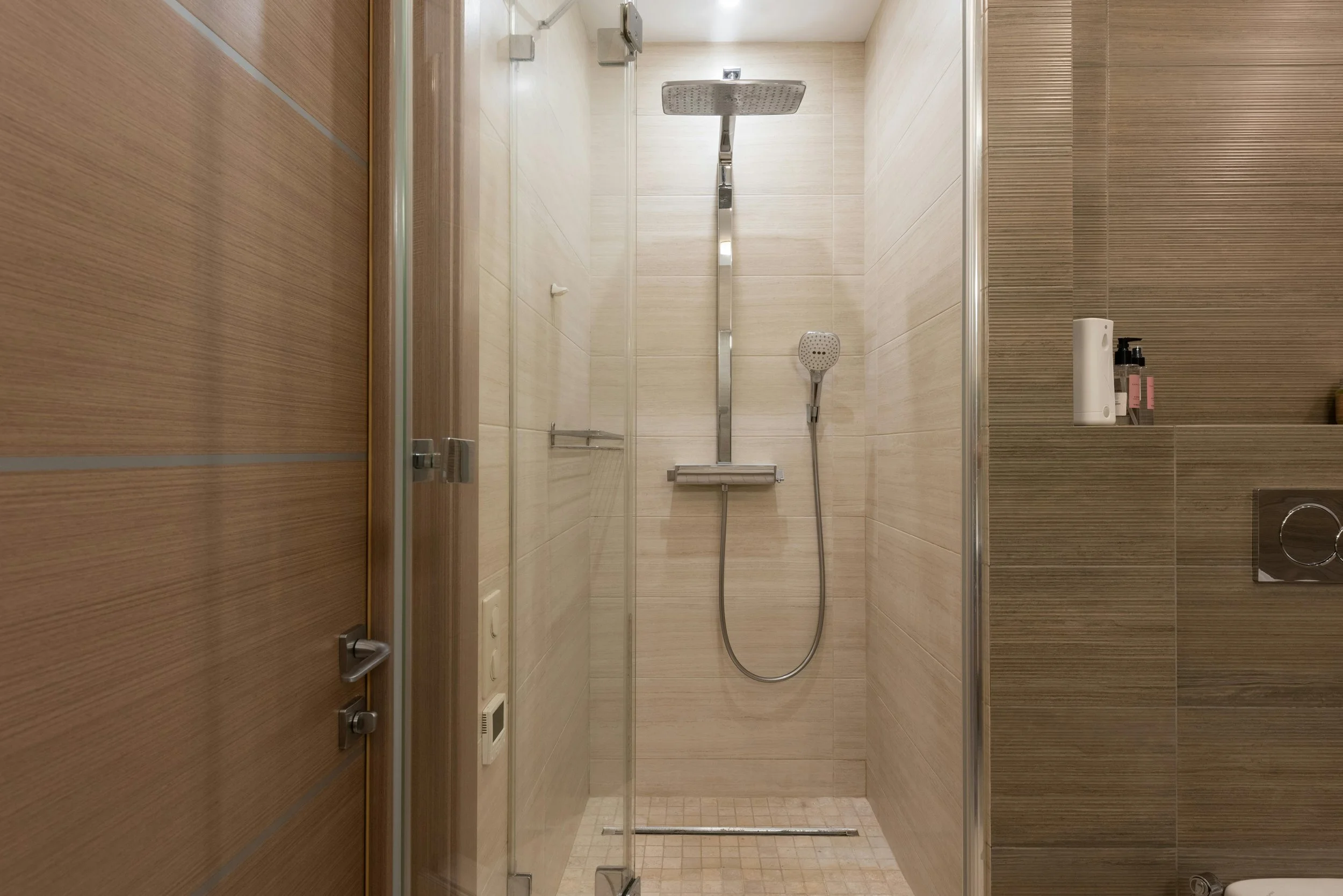 View of a modern, enclosed shower with beige and brown tiled walls, a stainless steel rainfall showerhead, a handheld shower, and a built-in shelf with toiletries.