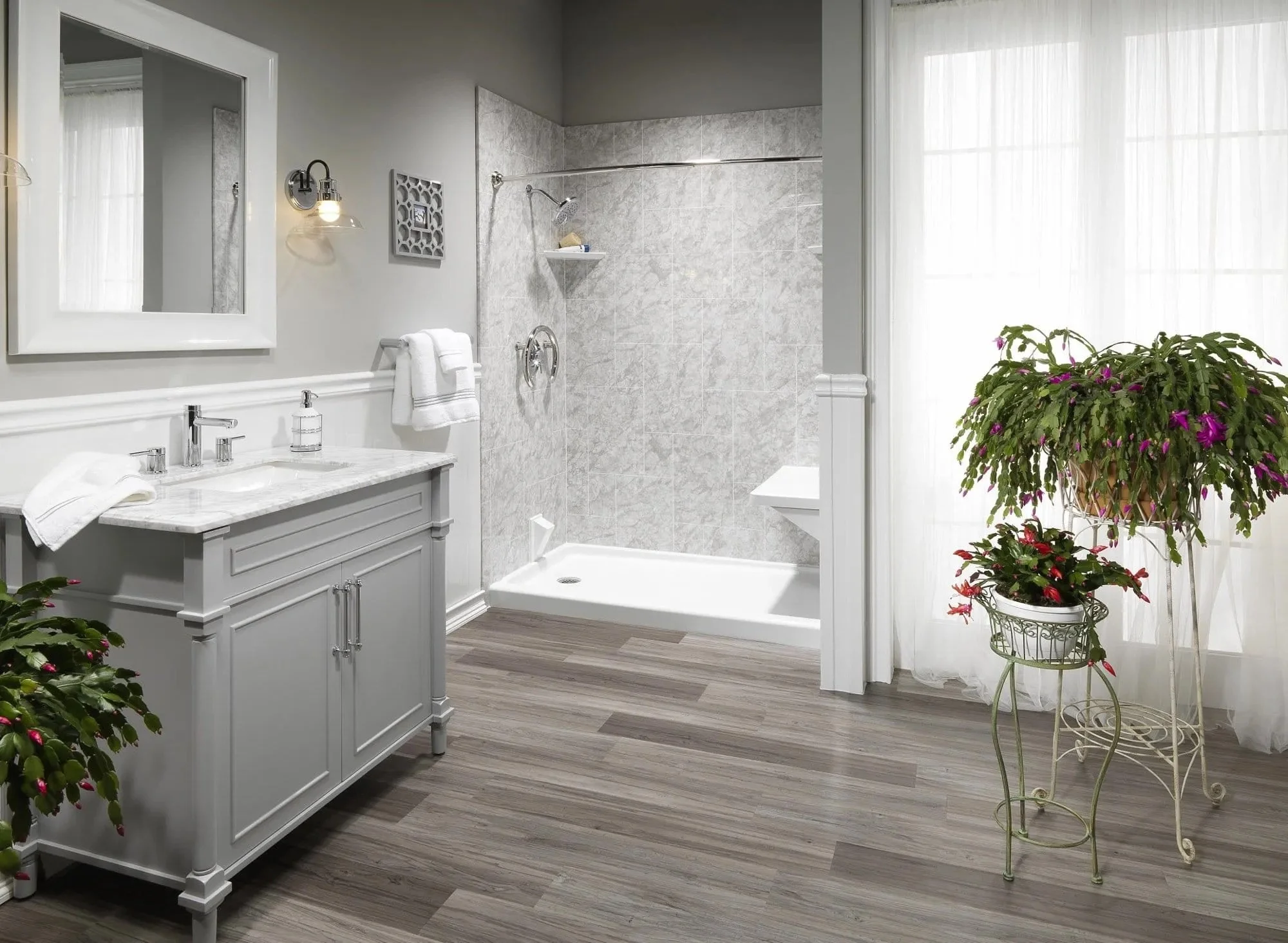 A clean, bright bathroom with a white vanity, marble countertop, and a mirror. There is a walk-in shower with gray tile, a large window with sheer curtains, and blooming potted plants on decorative plant stands.