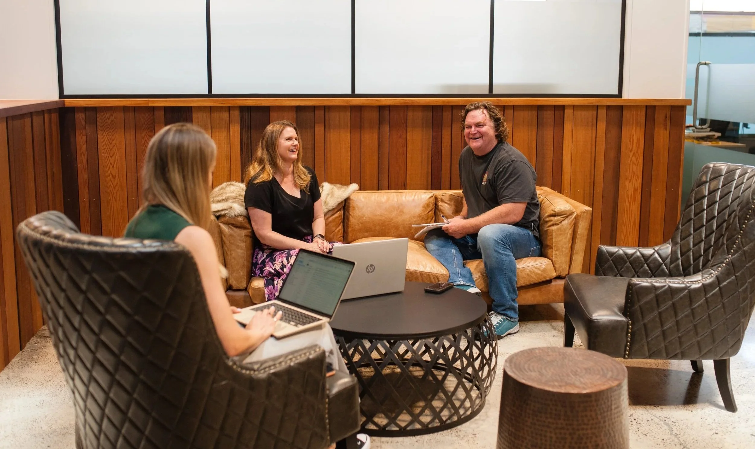 Three people sitting on a brown leather couch and chairs in a casual office setting, engaged in conversation and smiling.