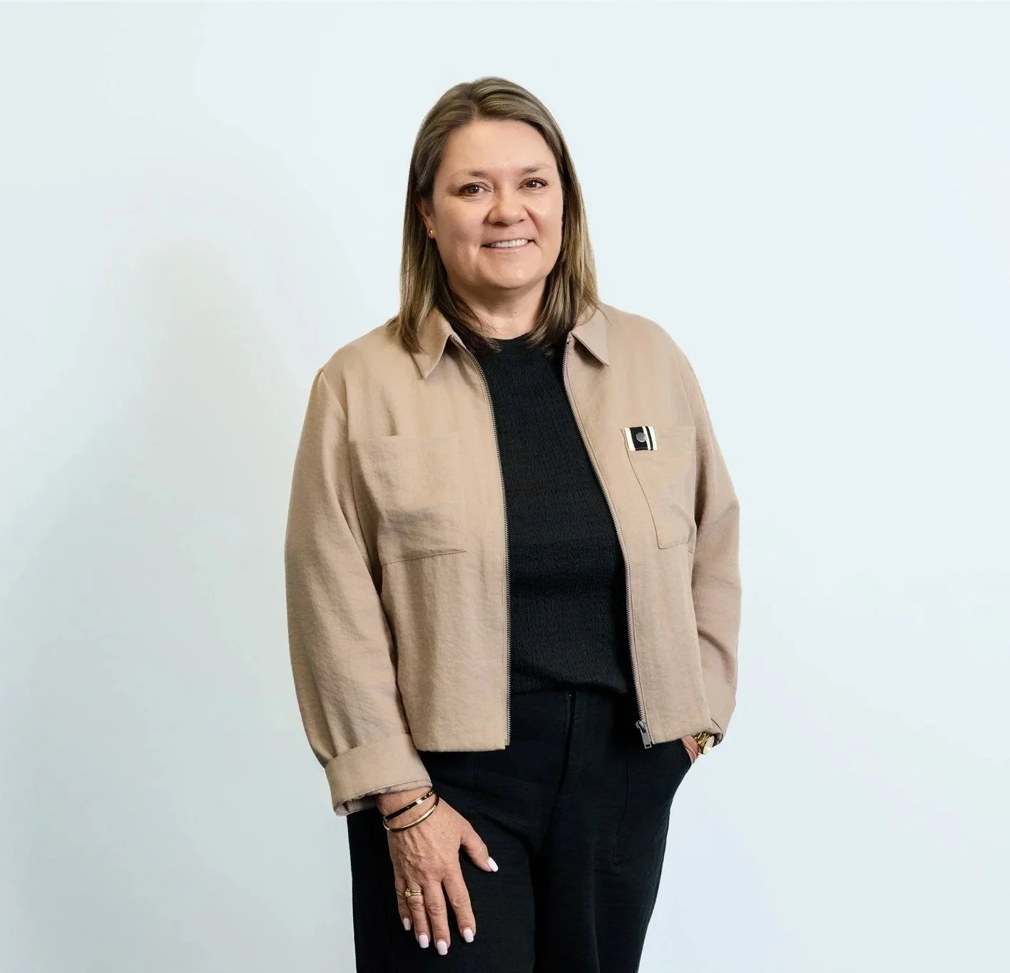 Woman in beige jacket and black shirt standing against a plain white background.