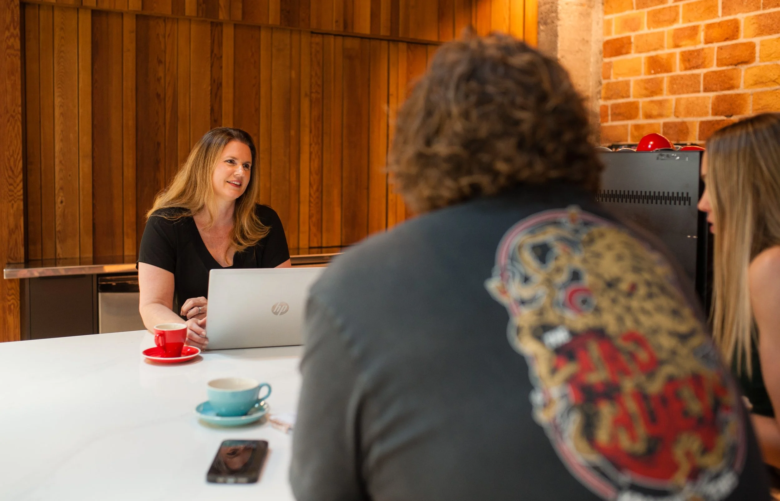 A woman sitting at a table with a laptop, talking to two people across from her in a cafe or meeting room with wood-paneled and brick walls.