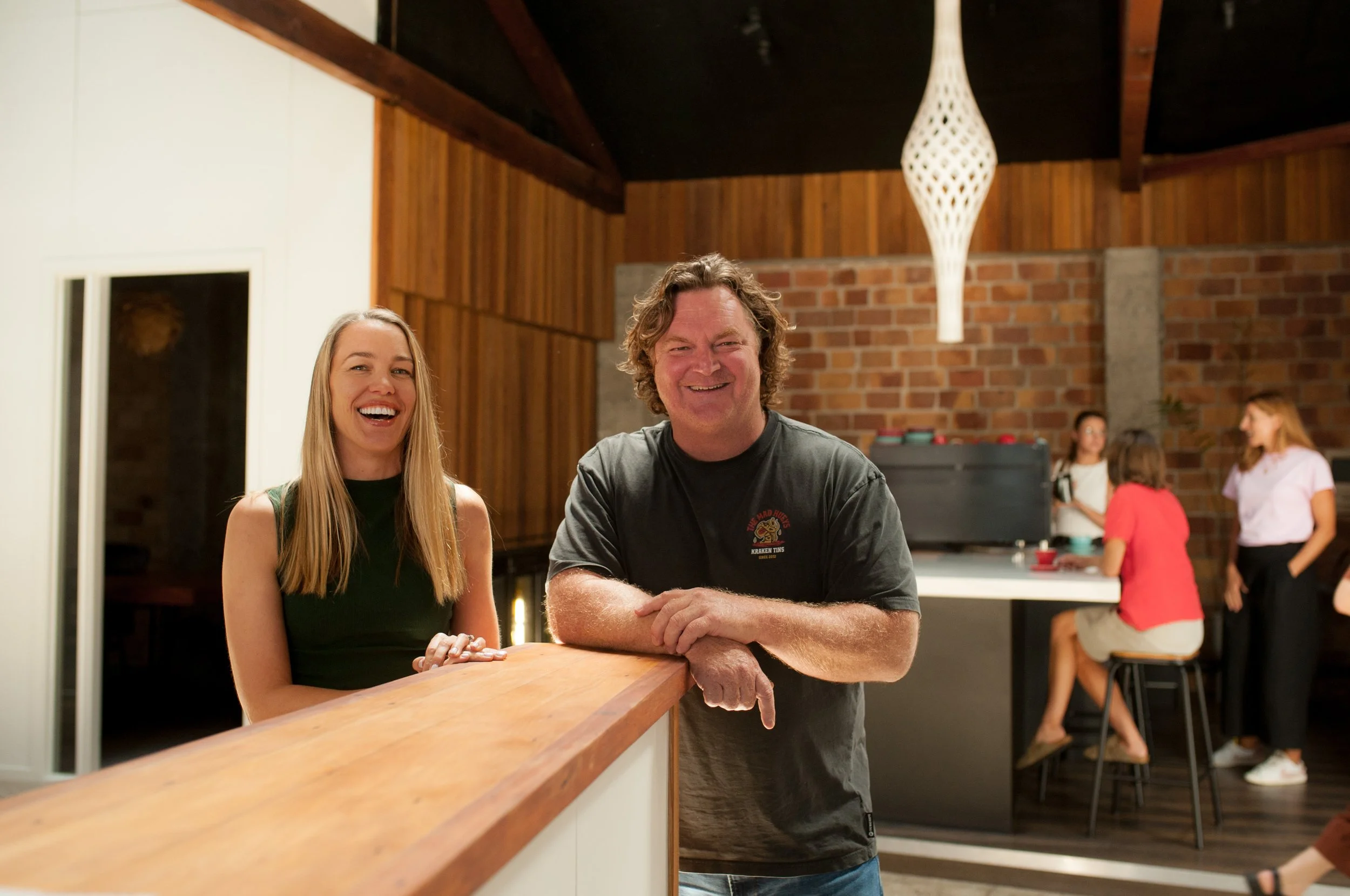 A man and a woman standing at a bar counter, smiling and laughing, with a group of women in the background near a kitchen counter in a cozy, modern room.