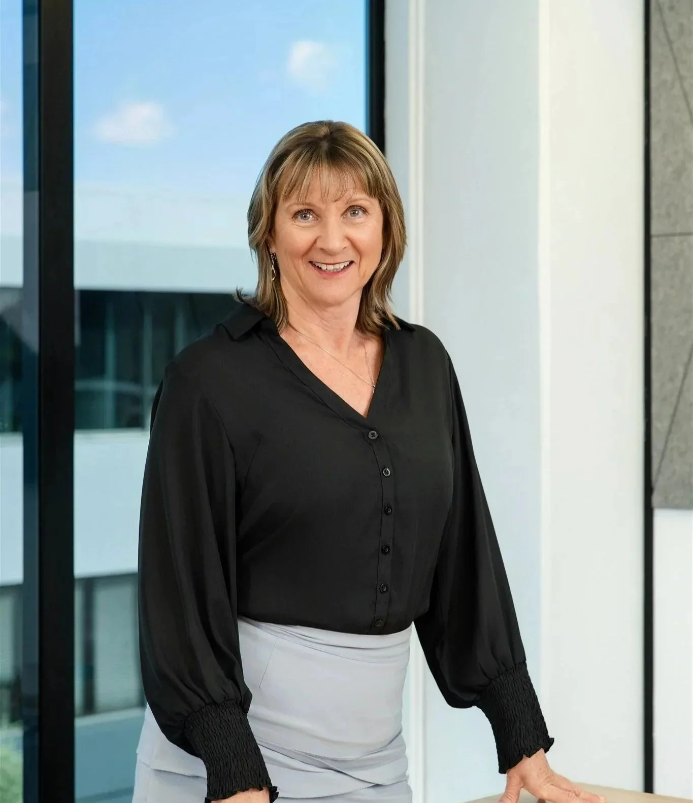 Smiling woman with light brown hair in a black blouse, standing near large windows, in an office setting.