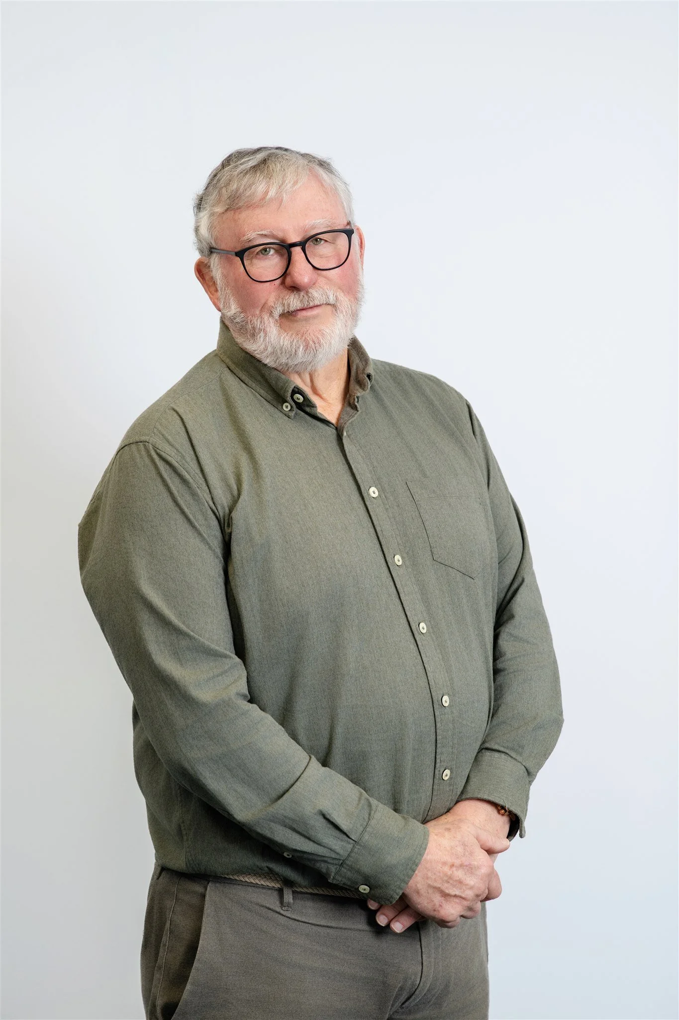 An older man with white hair, glasses, and a beard posing against a plain white background. He is wearing a green button-up shirt and brown pants.