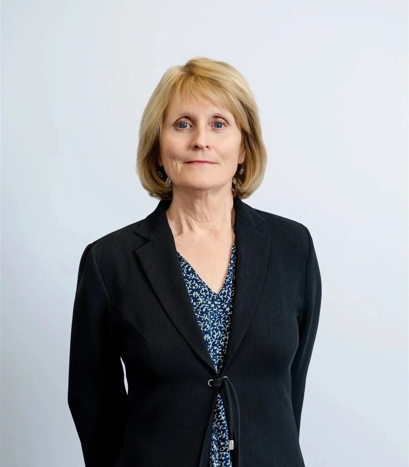 Portrait of a middle-aged woman with blonde hair, wearing a black blazer over a blue patterned blouse, standing against a plain light background.