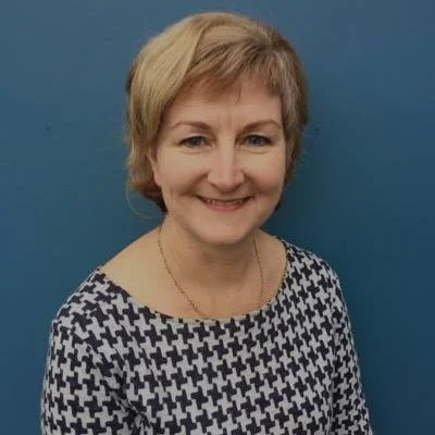 A smiling middle-aged woman with short blonde hair, wearing a patterned top, standing against a blue background.