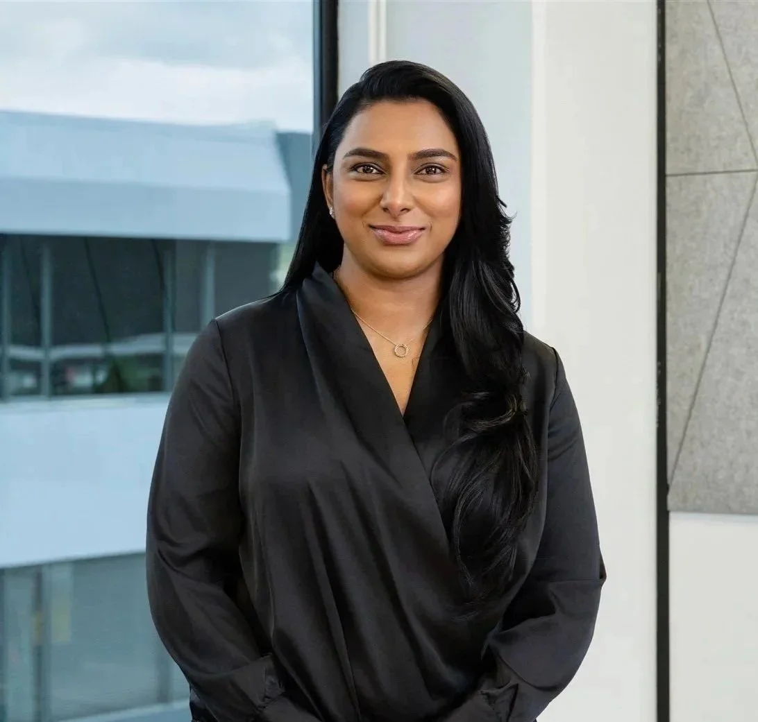 Portrait of a woman with long black hair, wearing a black blouse, standing in front of a modern office building window.