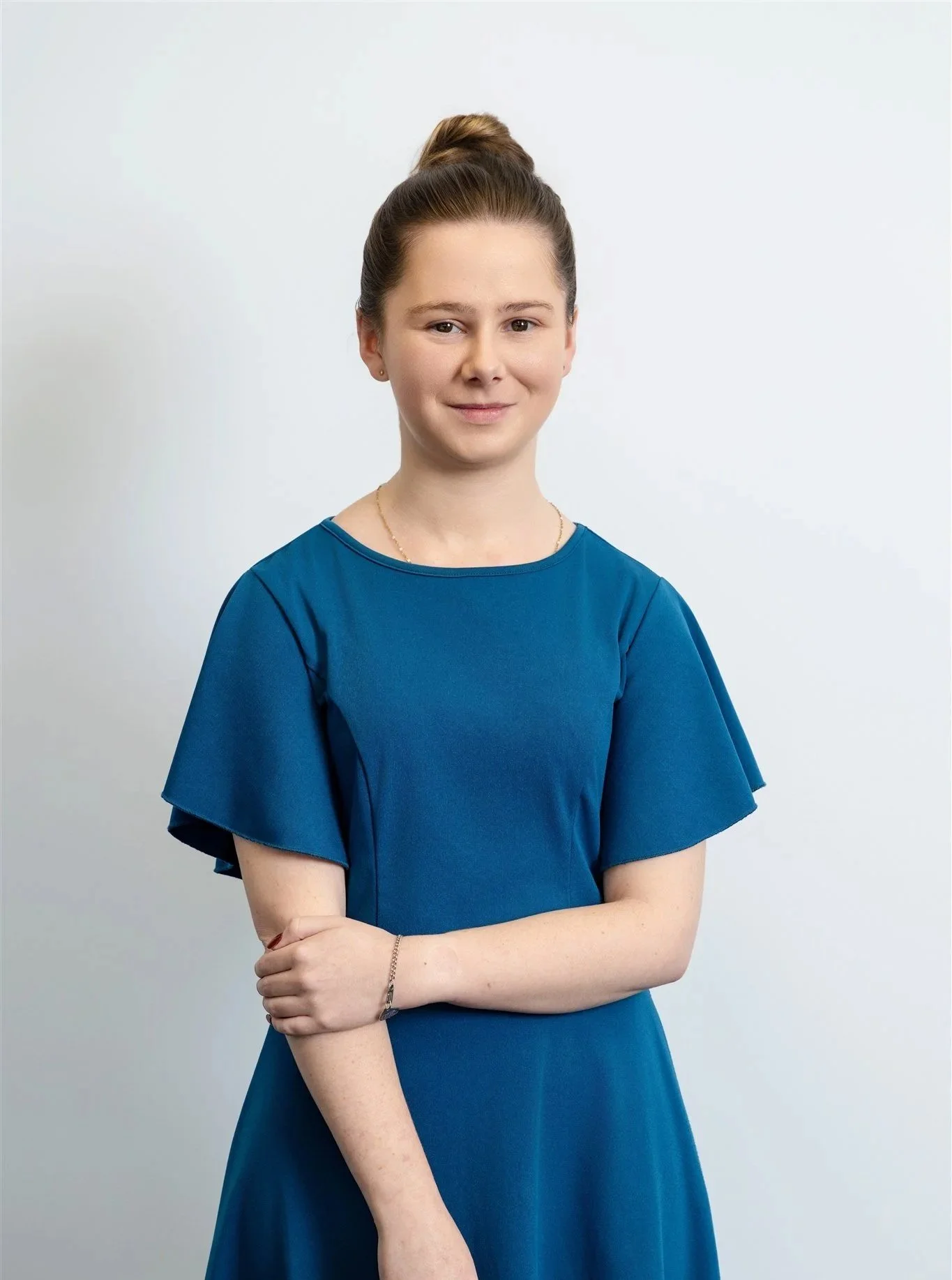 Portrait of a young woman with brown hair in a bun, wearing a blue dress, standing against a plain light background.