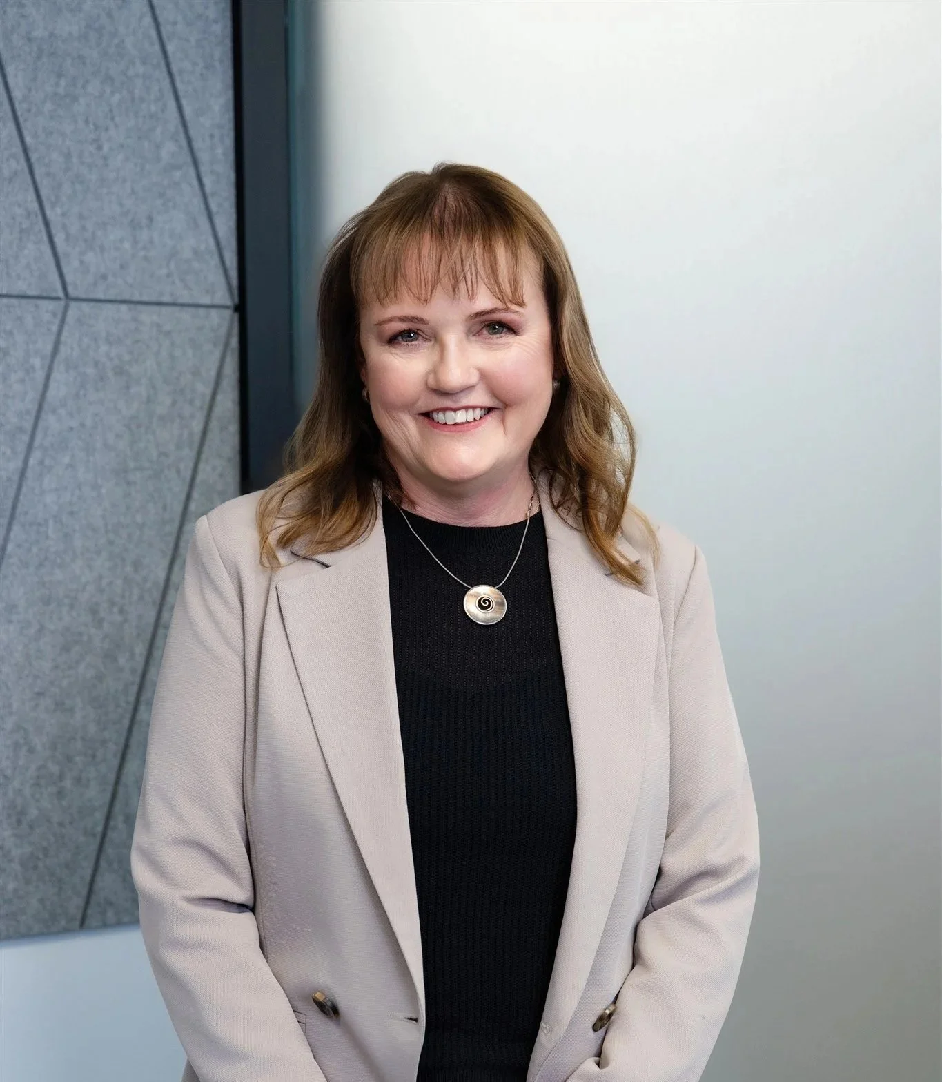 A woman with shoulder-length light brown hair and bangs, wearing a black top, light beige blazer, and a circular pendant necklace, standing in front of a geometric wall with a white background.