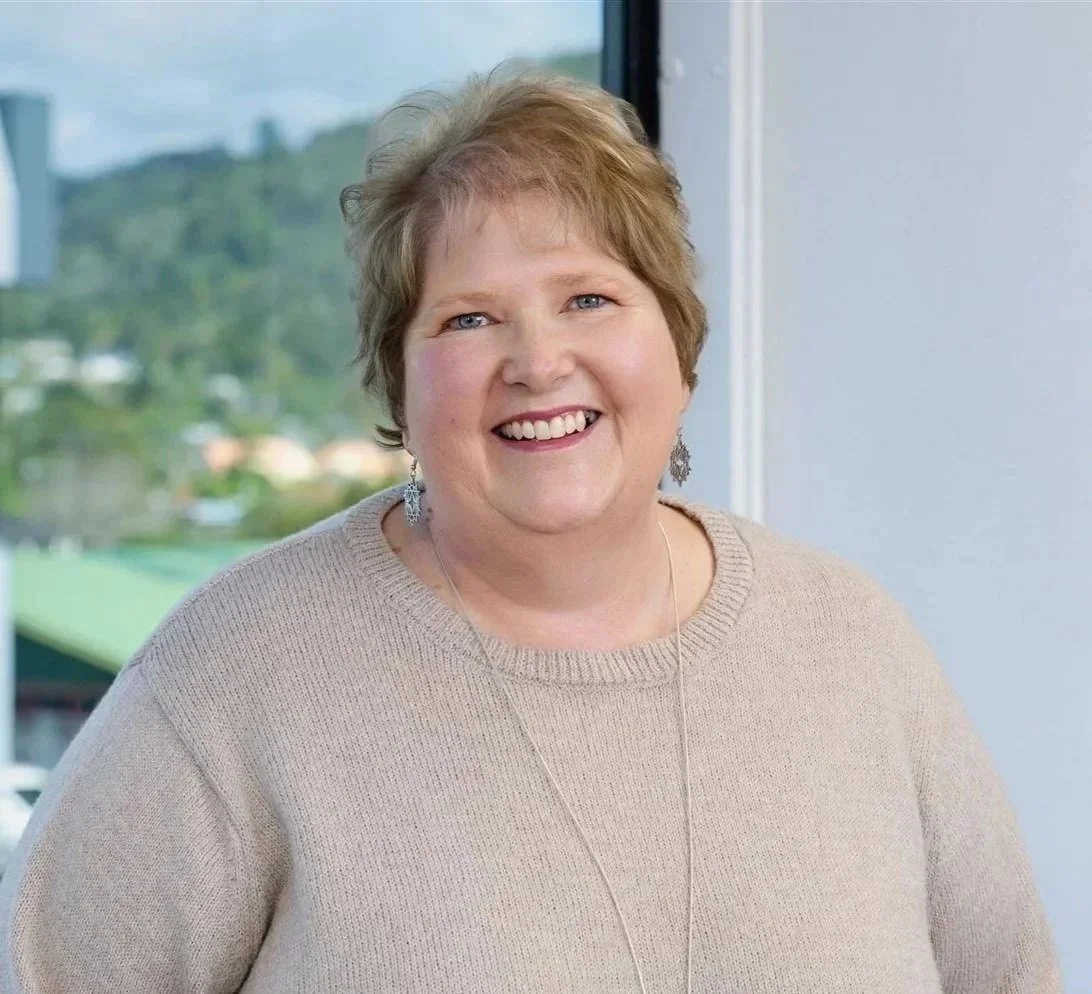A smiling middle-aged woman with short, light brown hair and blue eyes, wearing a beige sweater and dangling earrings, standing indoors by a window with a blurred outdoor landscape in the background.