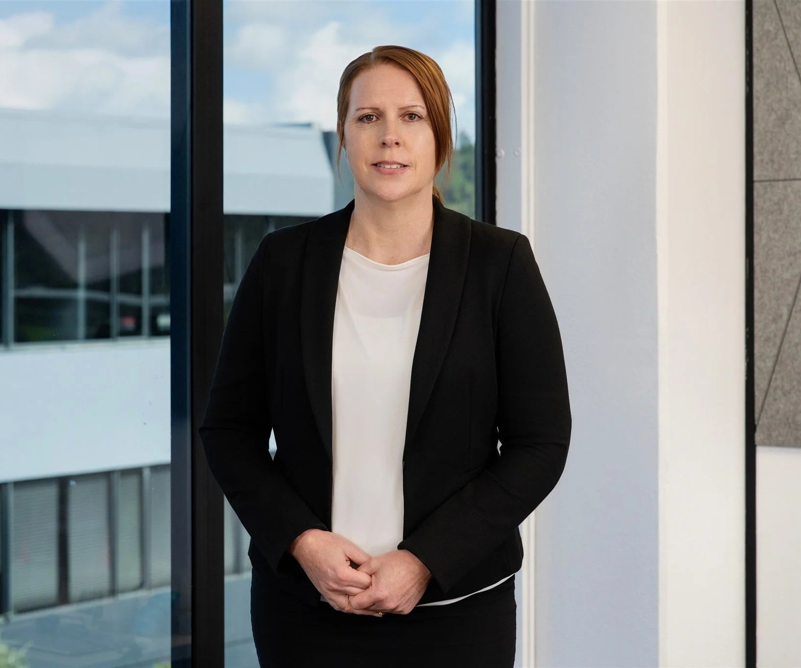 A woman with short red hair wearing a black blazer over a white top, standing indoors near large windows with a modern building visible outside.