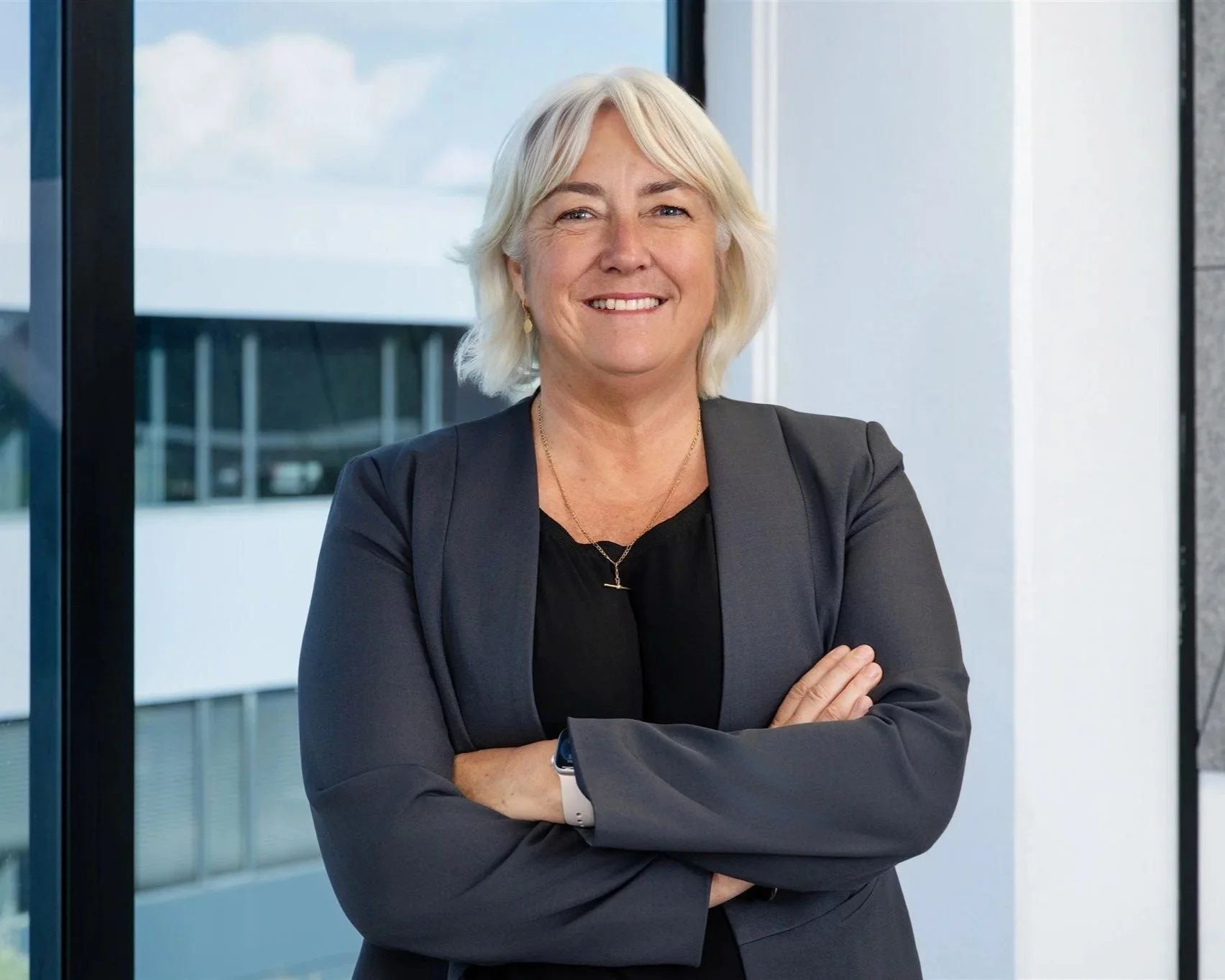 A smiling senior woman with blonde hair wearing a black top and grey blazer, standing with arms crossed in front of a large window in a modern office building.