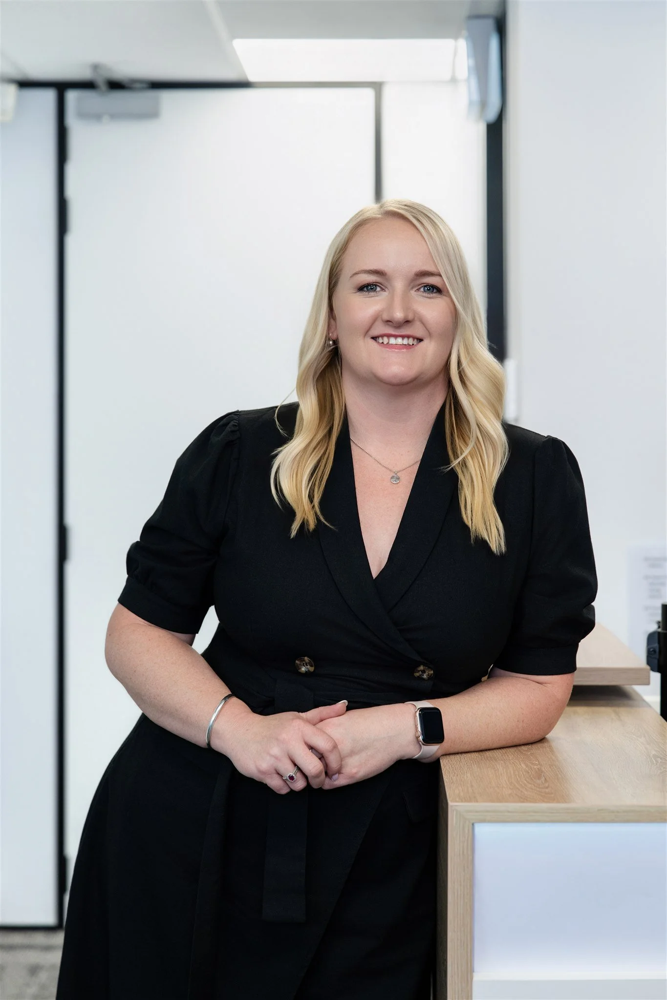 A smiling woman with blonde hair, wearing a black dress, standing next to a wooden desk in an office setting.