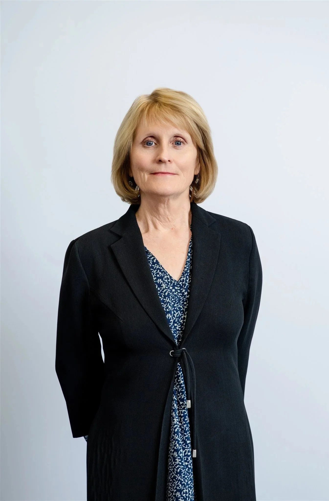 Photo of a middle-aged woman with blonde hair, wearing a dark blazer and a patterned blouse, standing against a plain light background.