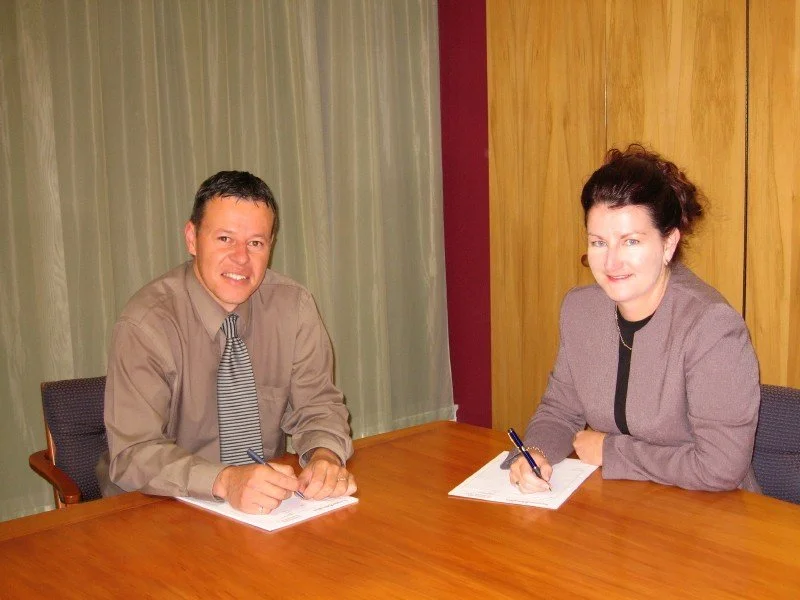 Two people, a man and a woman, sitting at a wooden table signing documents in a conference room.