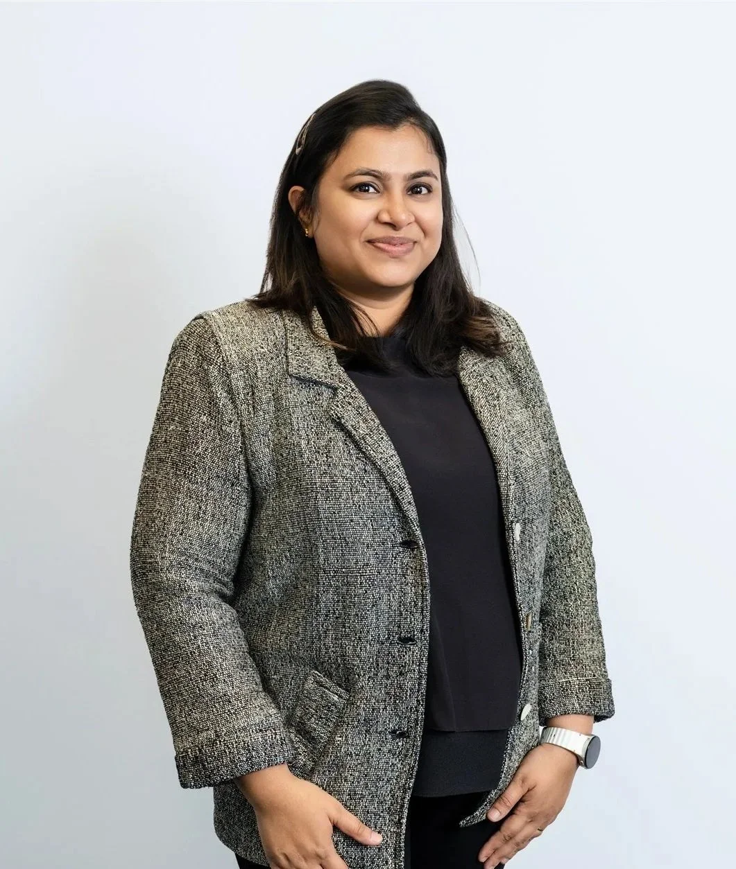 A woman with medium-length dark hair, wearing a black top, gray textured blazer, and a white wristwatch, standing against a plain white wall, smiling at the camera.