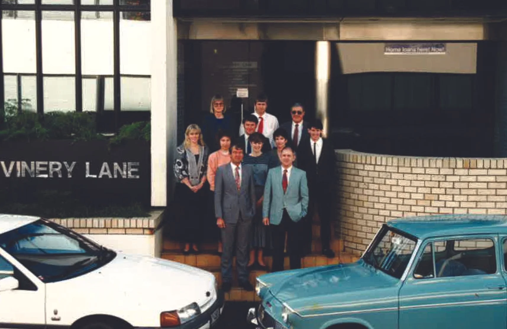 Group of people standing outside near a building with a sign that reads 'VINEY LAN' and two cars parked in front.