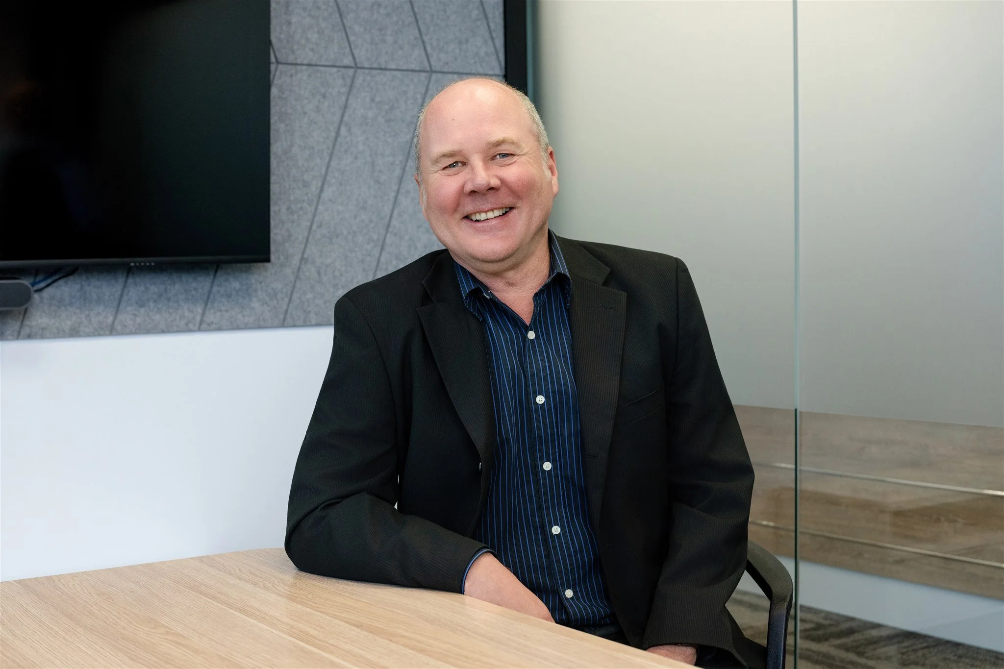 A smiling man in a black blazer and blue shirt sitting at a wooden table in a modern office setting.