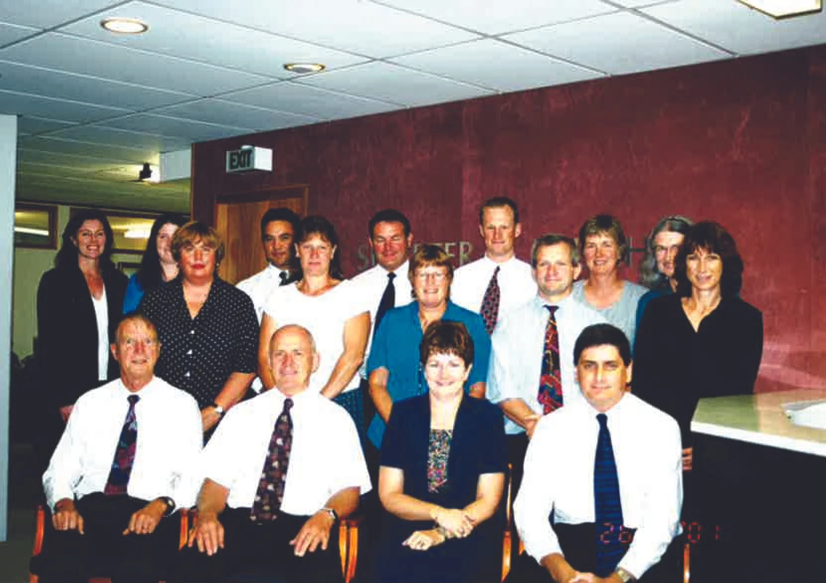 A group of 14 people, including men and women, gathered in a restaurant or banquet hall for a photo. They are dressed in business or semi-formal attire, with some men wearing white shirts and ties, and women in blouses and jackets. The background shows a red wall with some signage and a door. The group is smiling and posing for the picture.