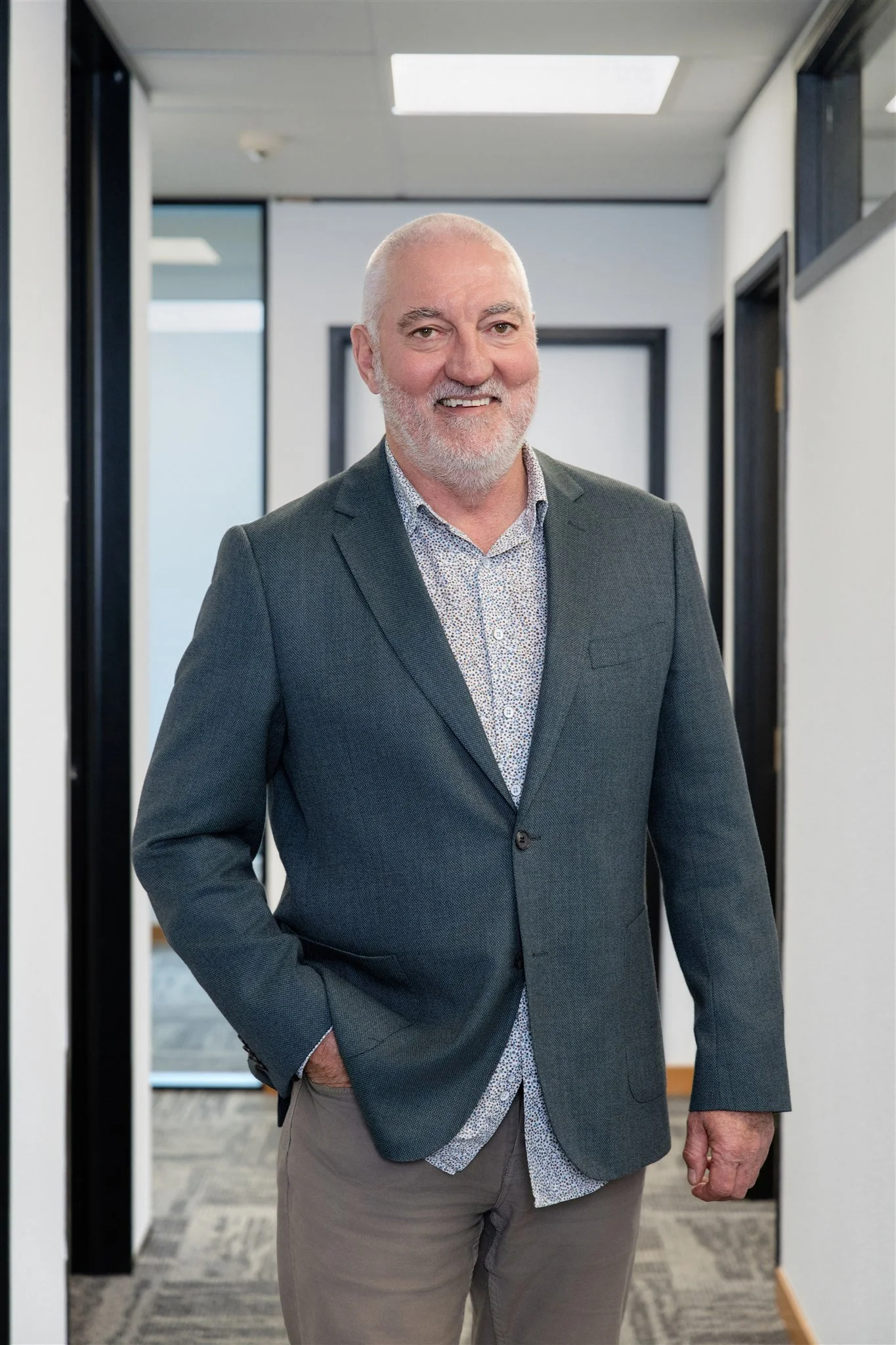 A middle-aged man with a beard and short gray hair wearing a gray blazer and patterned shirt, standing in an office hallway.