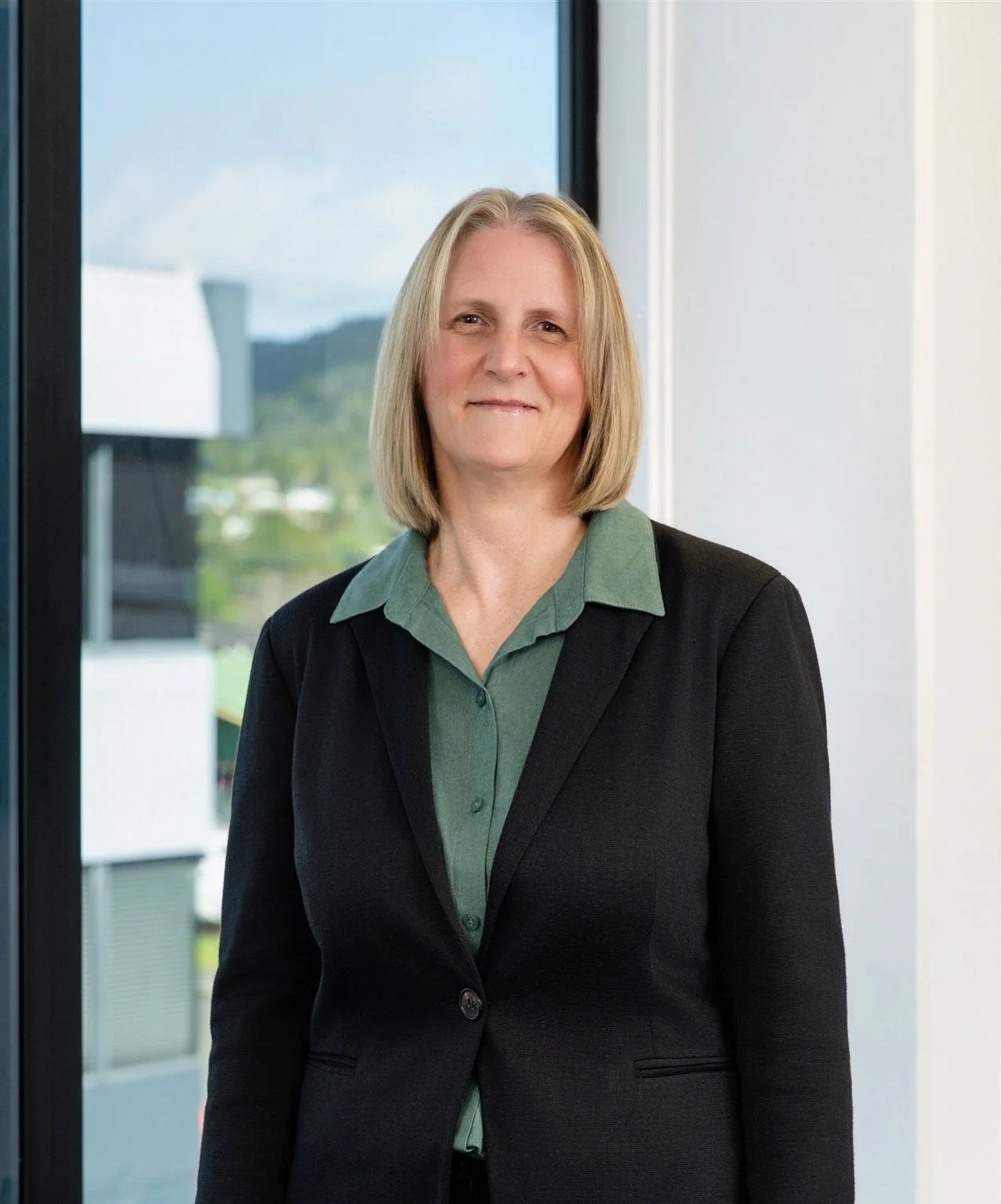 A middle-aged woman with shoulder-length blonde hair, wearing a dark blazer over a green button-up shirt, standing inside near a large window with a view of a cityscape and mountains in the background.