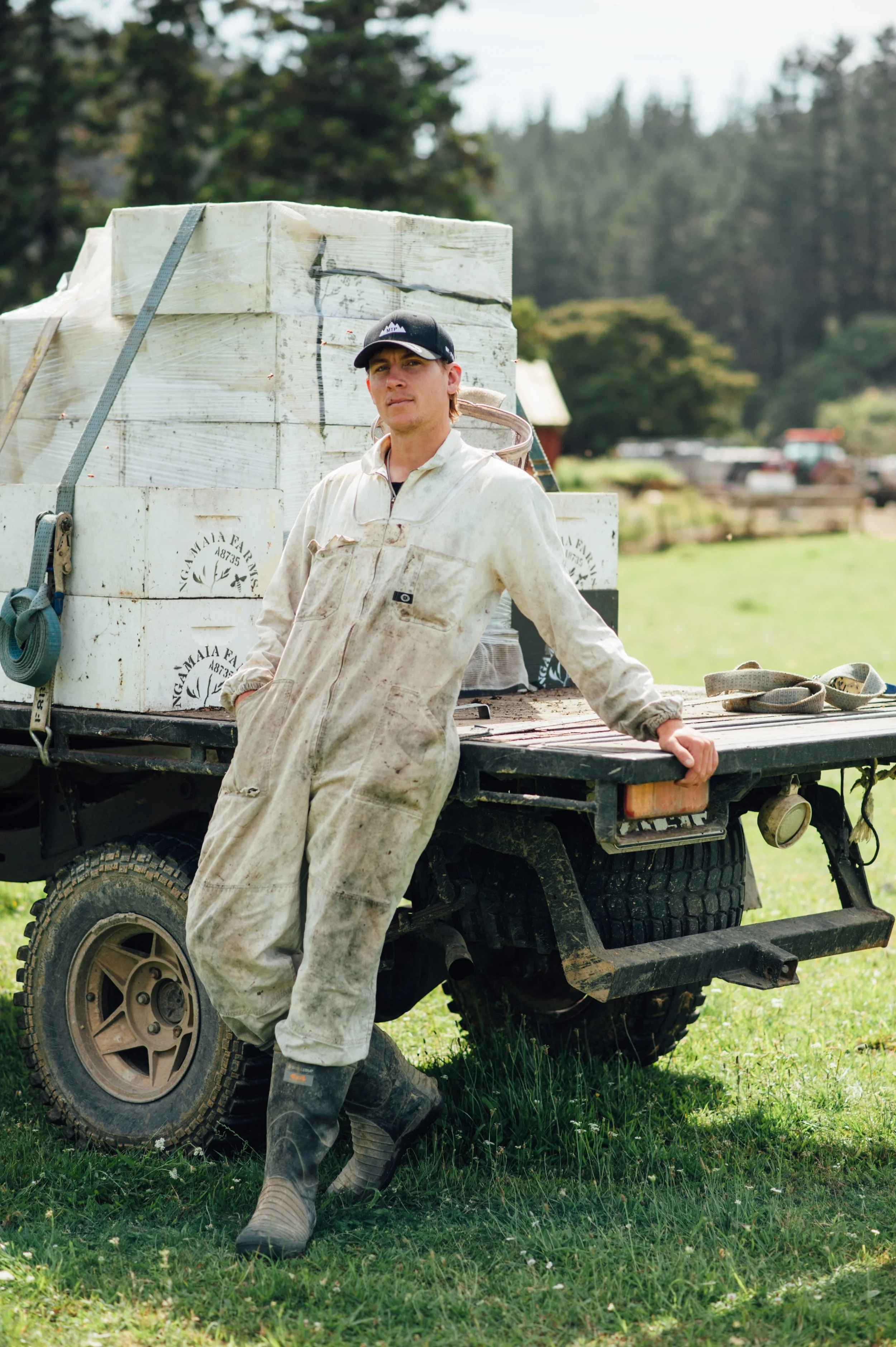 Young man in work overalls and boots leaning against a flatbed truck with boxes in outdoor rural setting.
