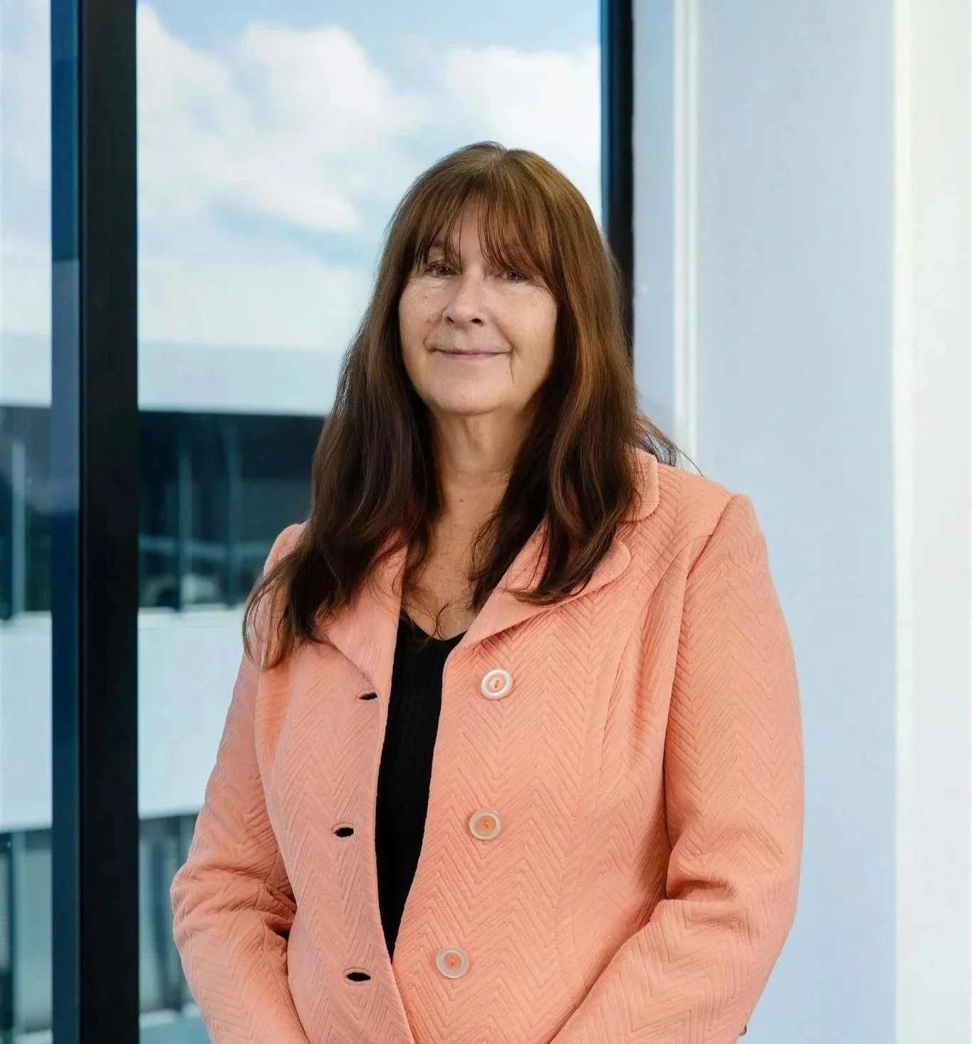 A middle-aged woman with brown hair and bangs, wearing a peach-colored blazer over a black top, standing indoors with large windows behind her showing a cloudy sky and building exterior.