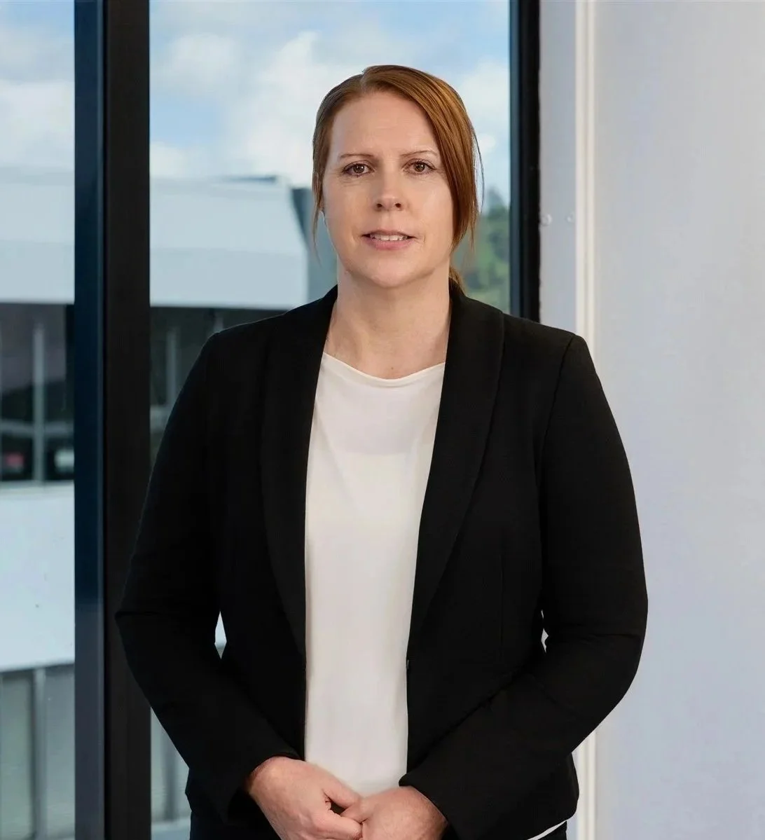 A woman with short red hair wearing a black blazer over a white top, standing in front of a large window with a view of a modern building outside.
