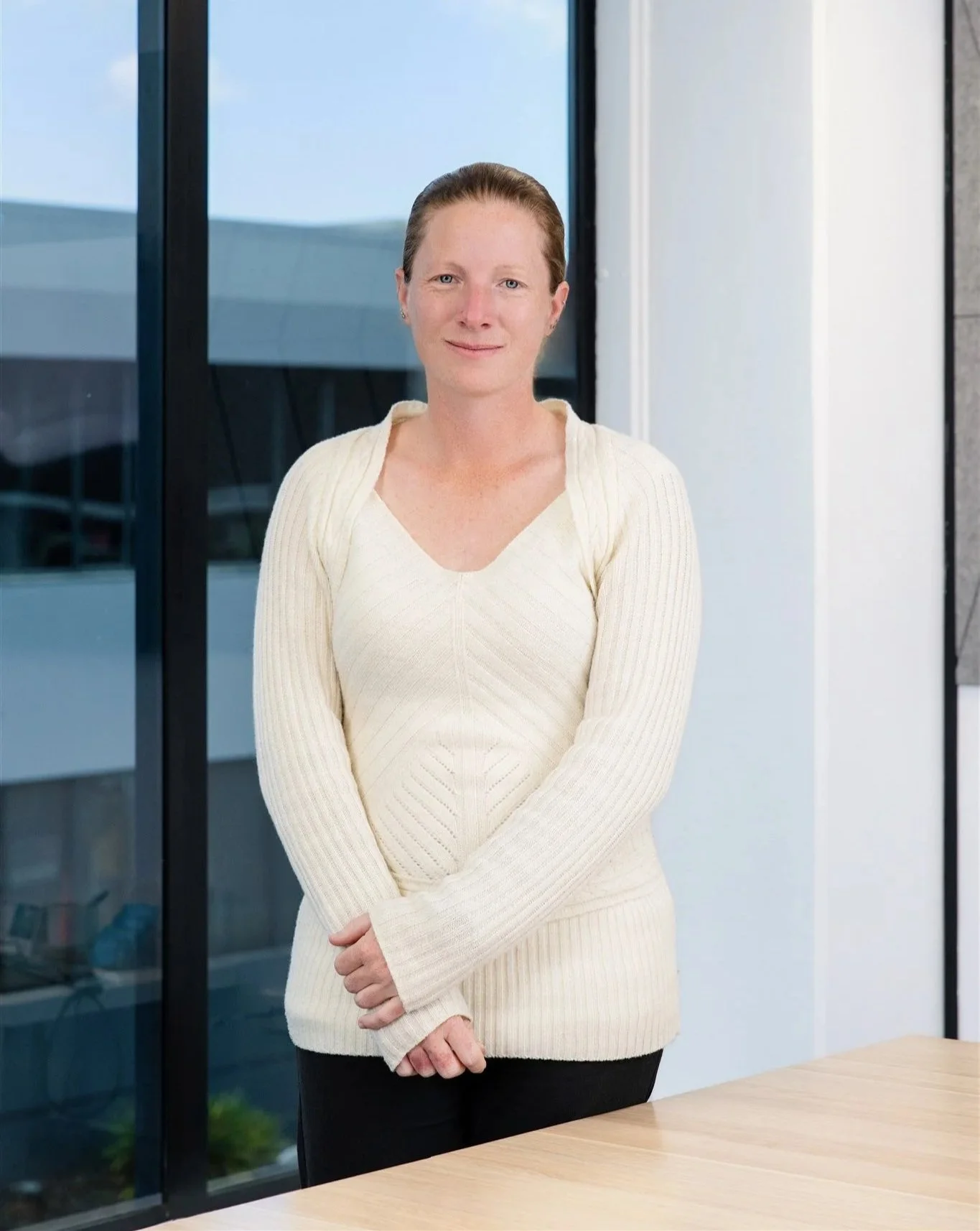 A woman with light skin and red hair tied back, wearing a cream-colored sweater, standing indoors near large glass windows with an urban cityscape in the background.