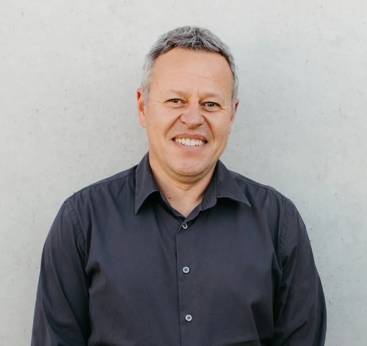 Smiling middle-aged man with gray hair wearing a black button-up shirt standing outdoors against a plain light-colored wall.