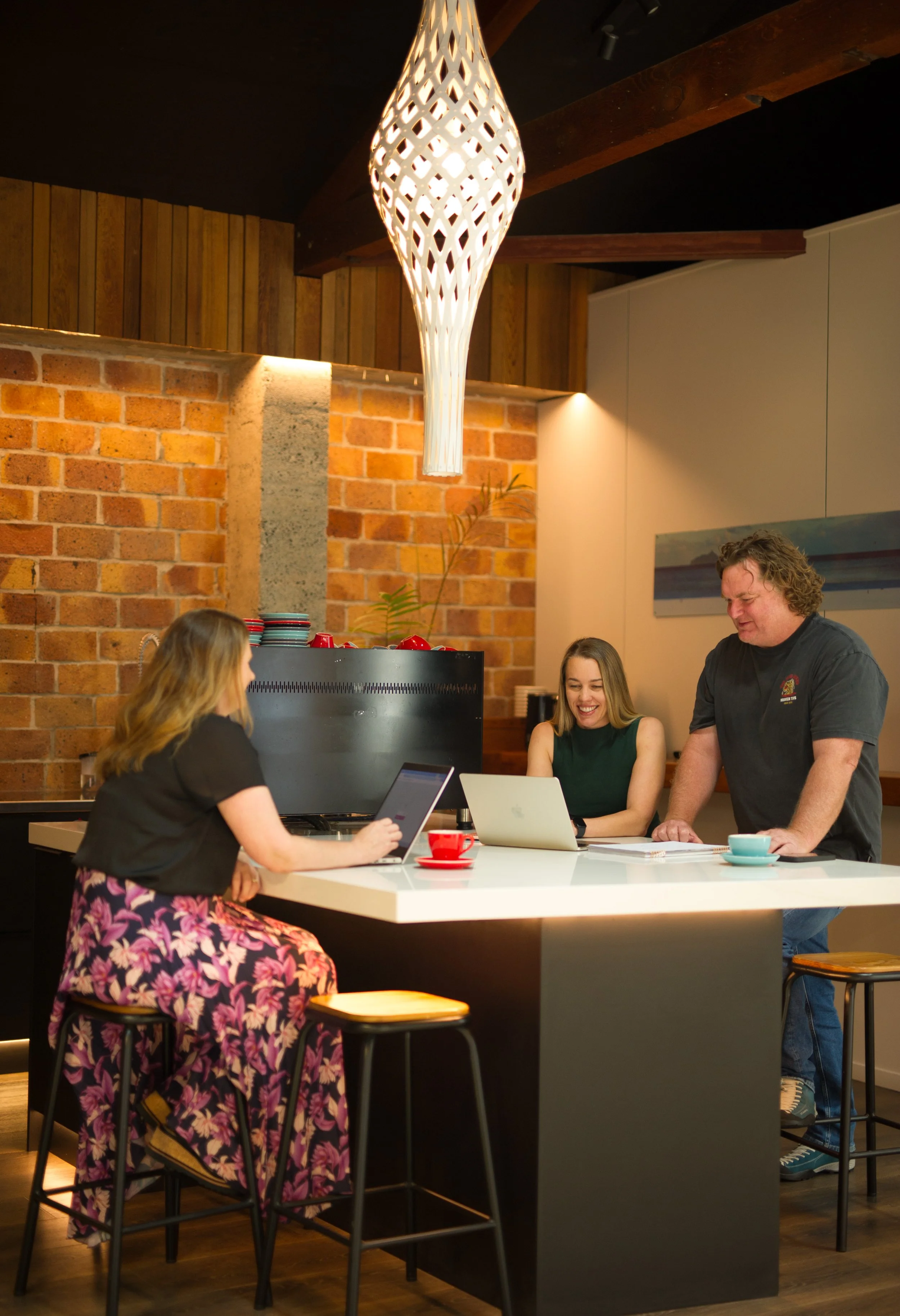 Three people sit at a kitchen bench at BigFish offices, with their laptops in front of them. They are smiling.