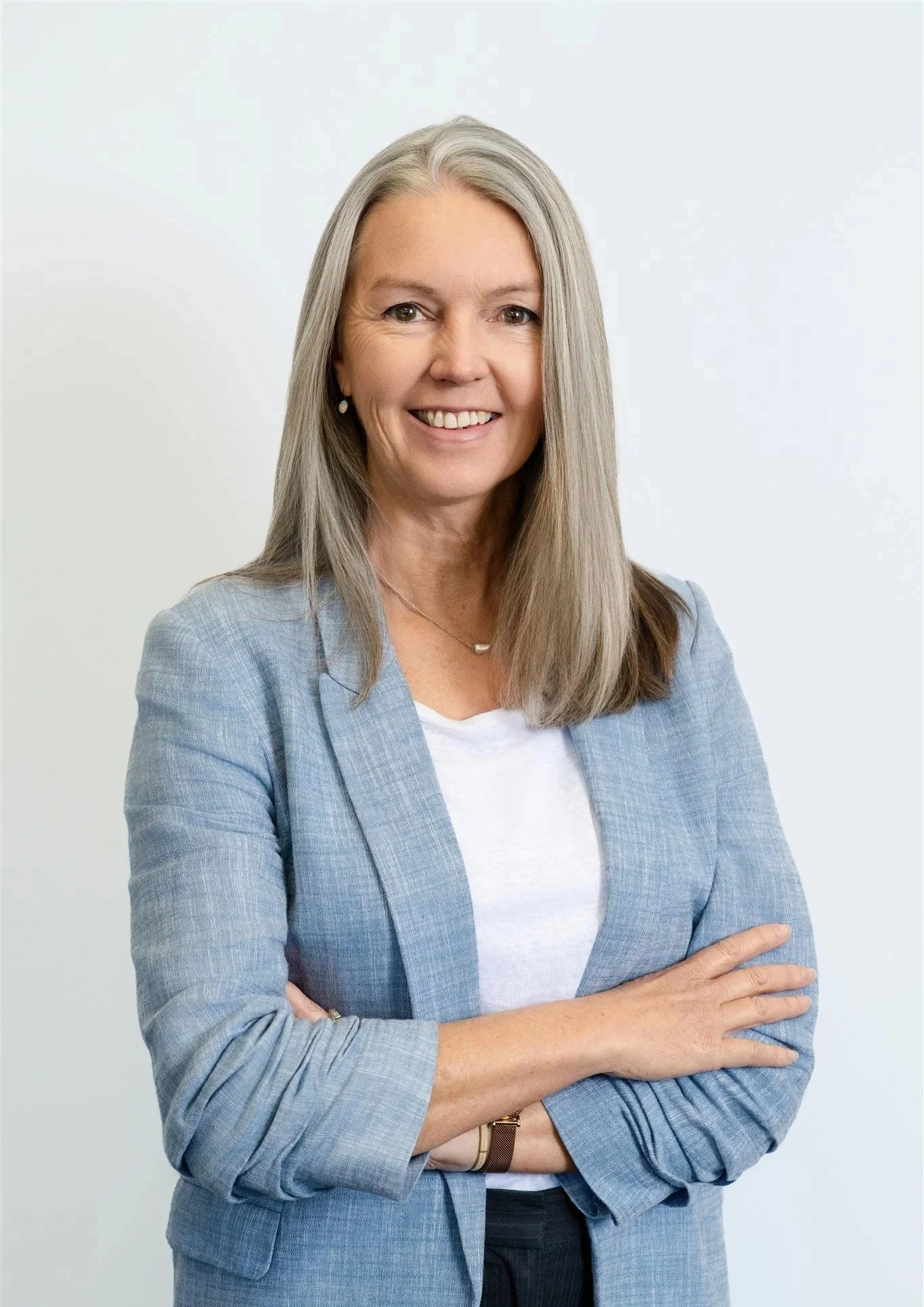 A middle-aged woman with long gray hair, smiling, wearing a light blue blazer over a white shirt, standing against a plain white background.