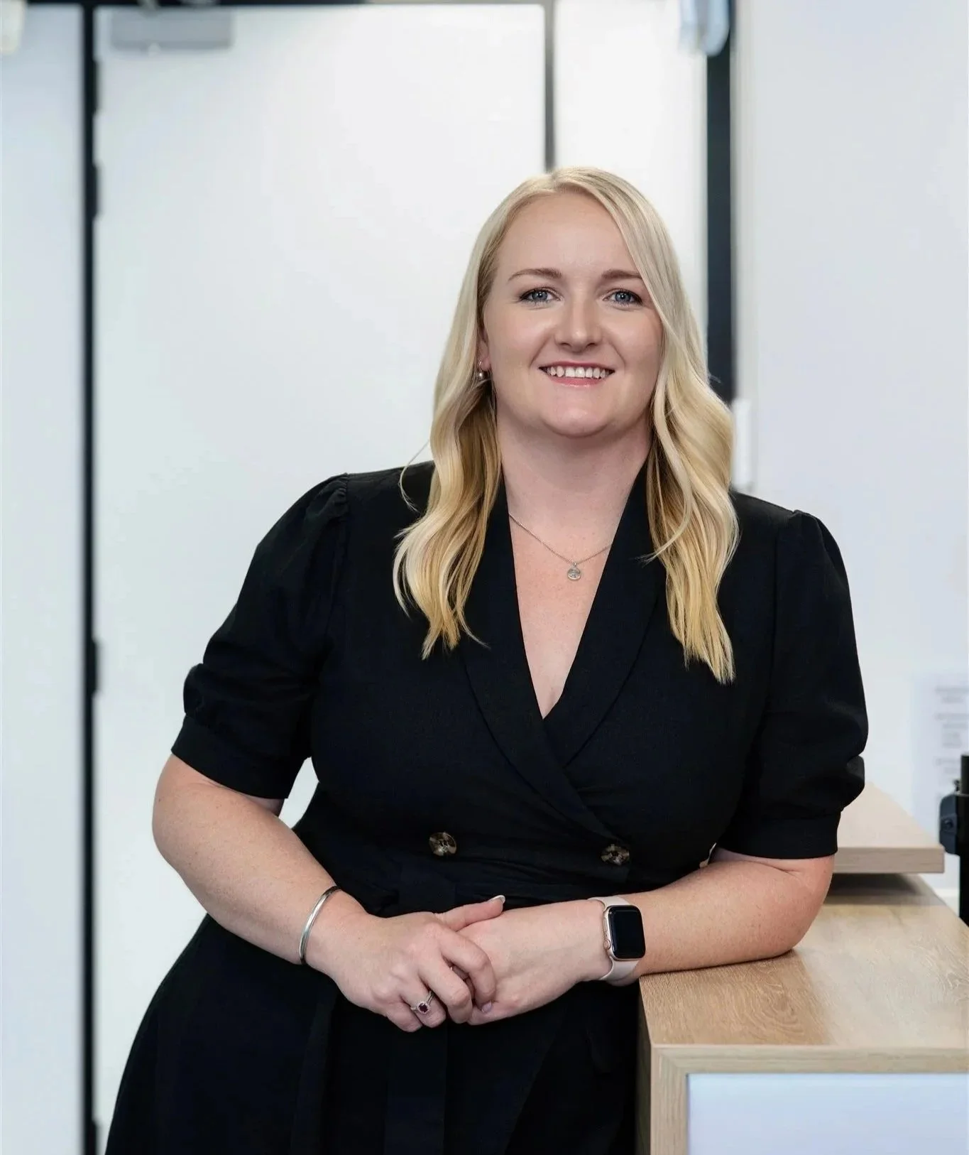 A woman with blonde hair wearing a black blazer, leaning on a wooden counter in a modern office setting.