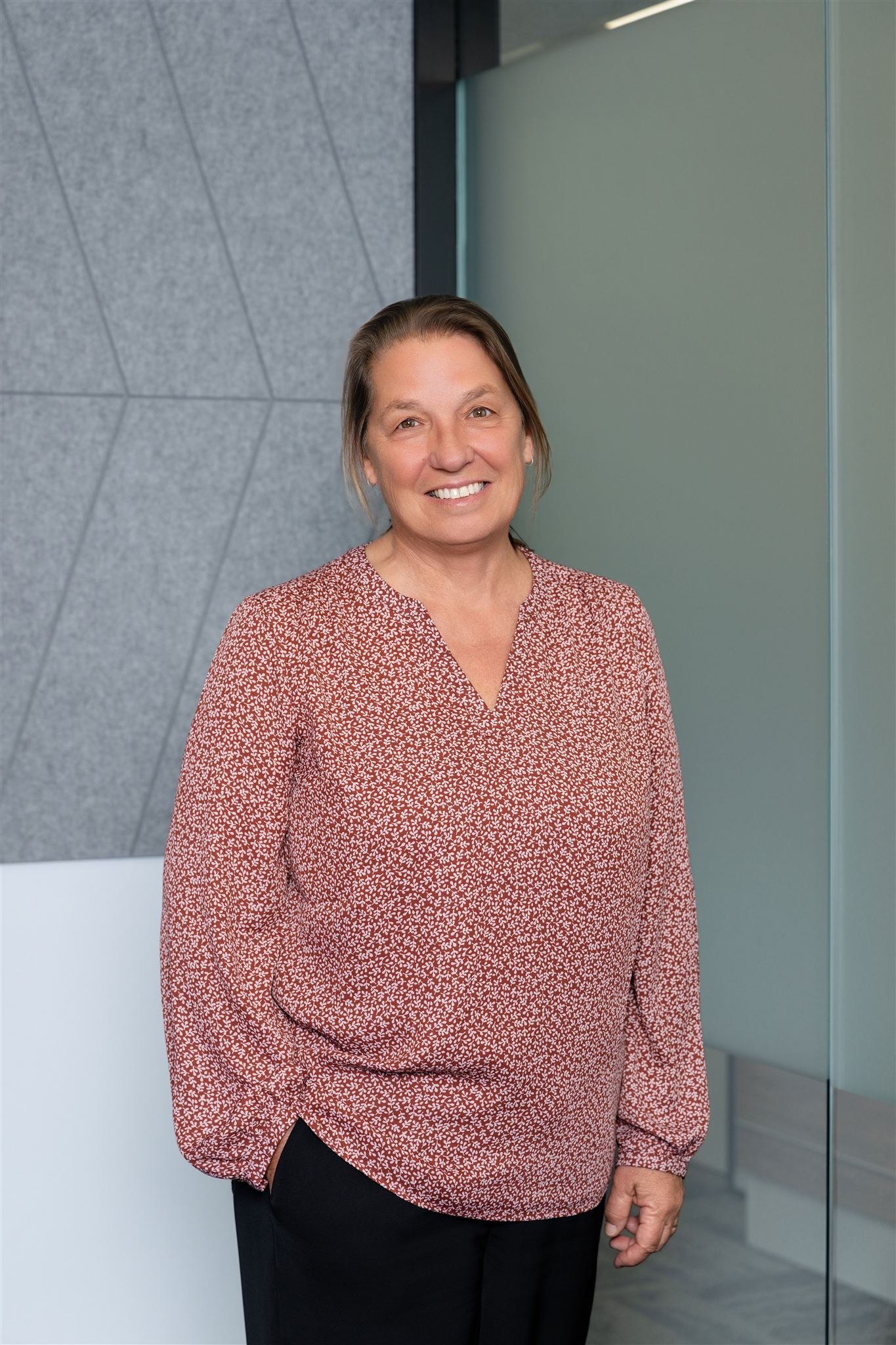 A woman with brown hair wearing a red patterned blouse and black pants standing indoors against a gray wall and frosted glass panel, smiling at the camera.