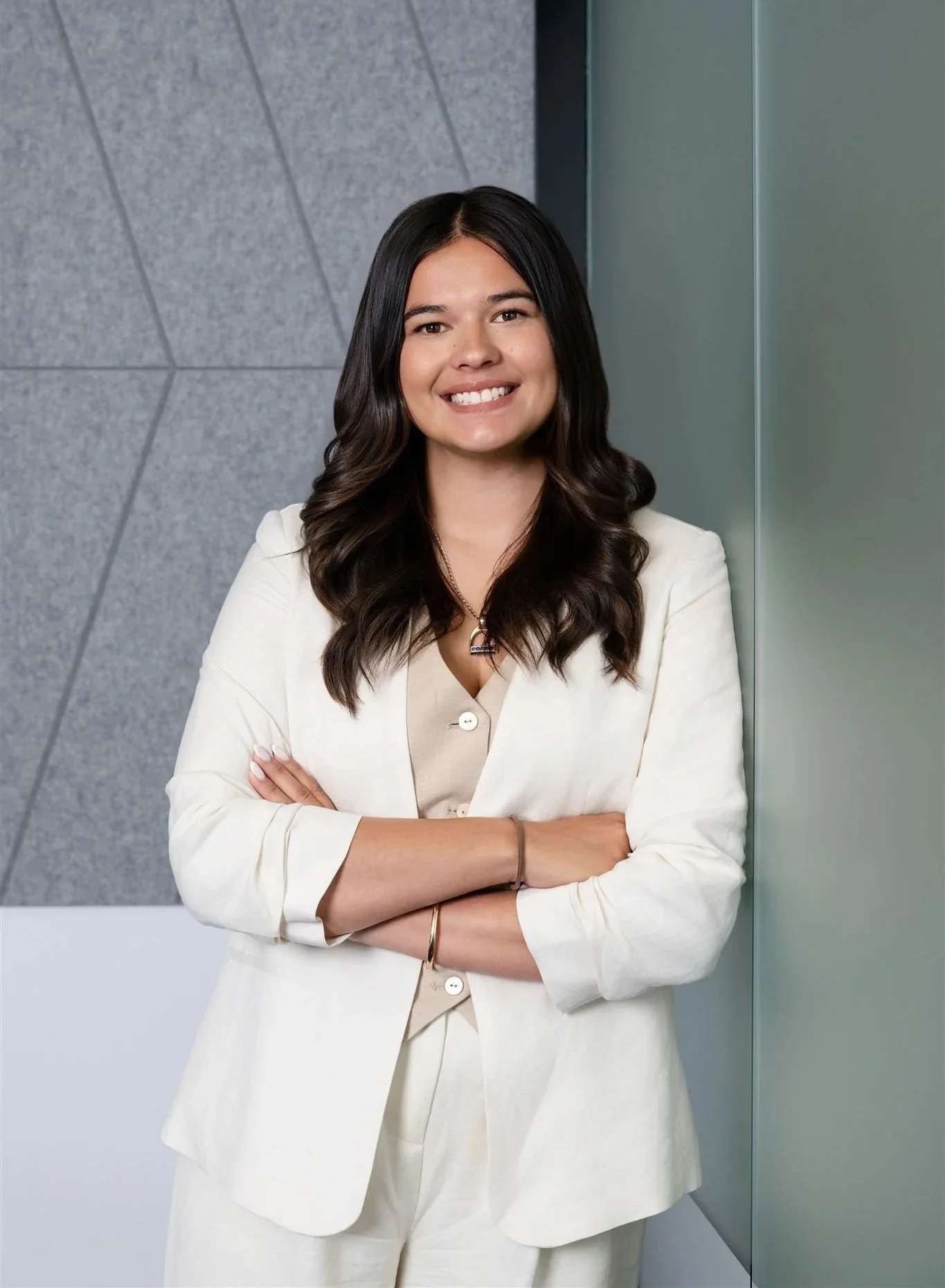 Portrait of a smiling woman with long dark hair, wearing a cream-colored blazer over a beige top, standing with arms crossed in front of a modern gray and green wall.