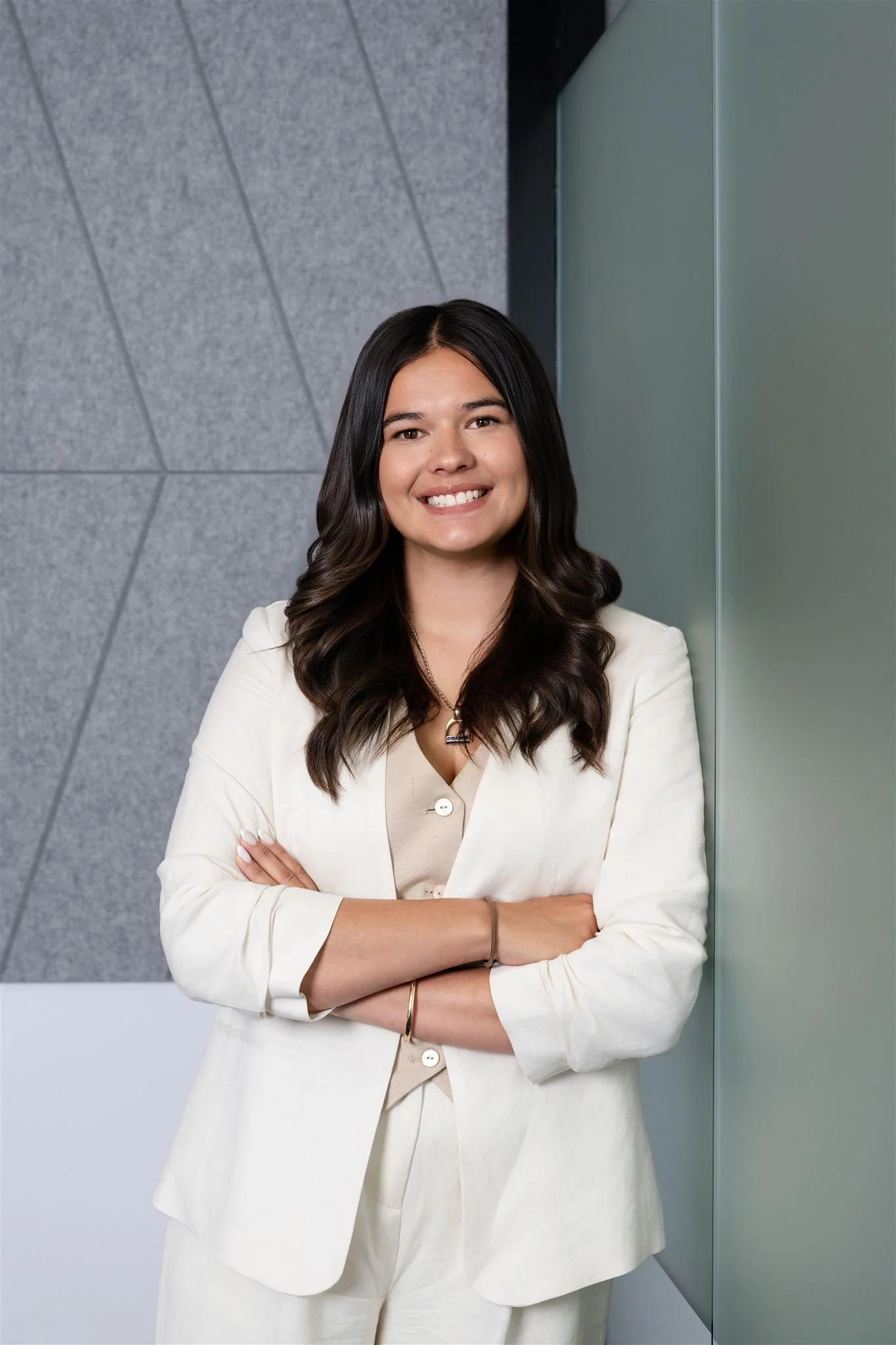 Portrait of a woman with dark wavy hair smiling, standing with crossed arms against a modern building wall.