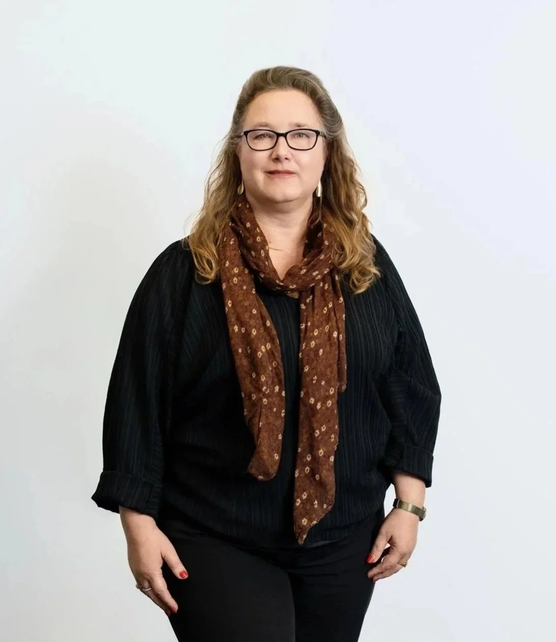 A woman with glasses and wavy, shoulder-length hair, wearing a black top, a brown patterned scarf, and black pants, standing against a plain white wall.