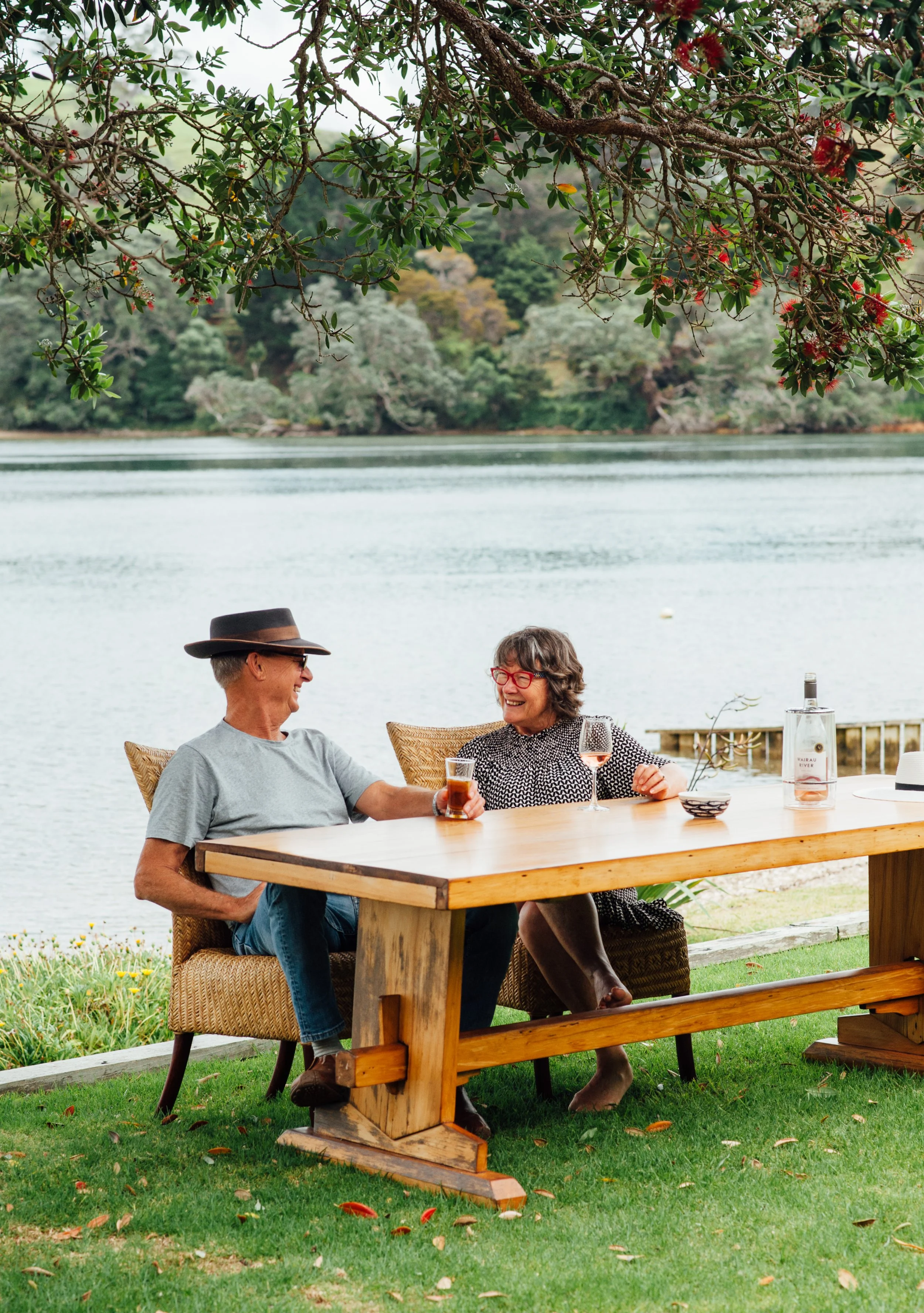 Two older adults, a man and a woman, sitting at a wooden table outdoors by a lake, smiling and enjoying drinks, with trees in the background.