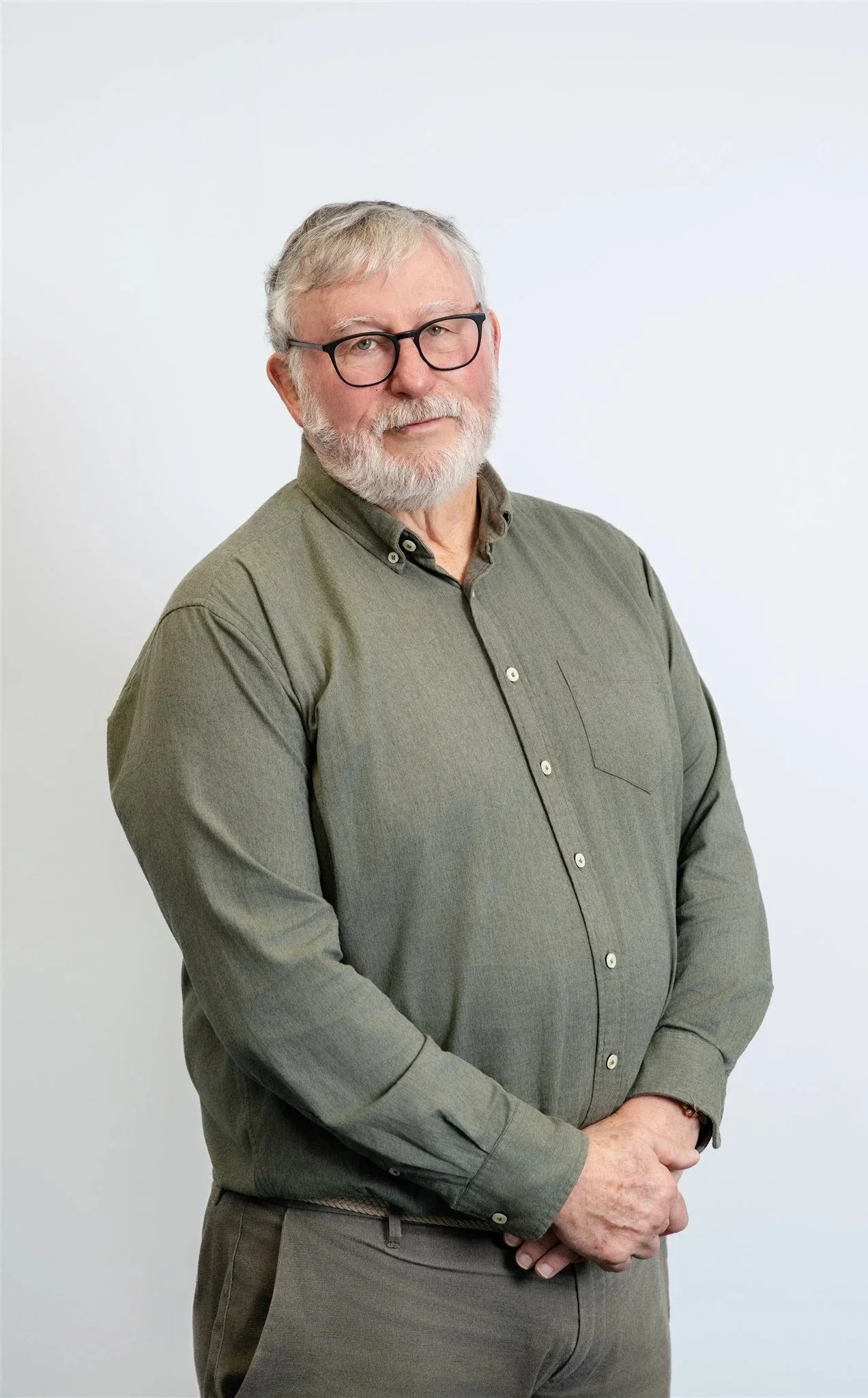 Portrait of an older man with glasses and a beard, wearing a green button-up shirt, standing against a plain white background.