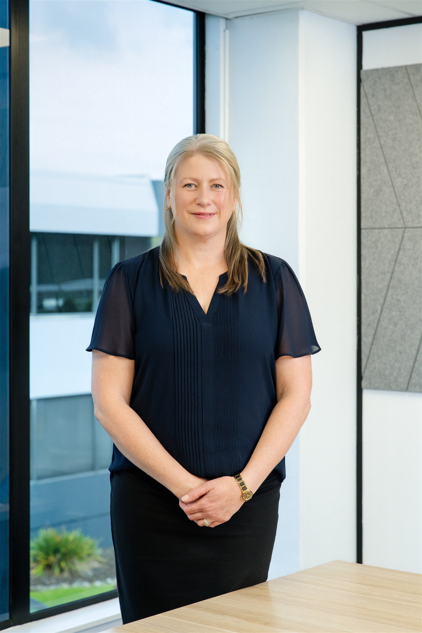 A woman with blonde hair, wearing a navy blouse and black skirt, standing in an office with large windows and modern interior design.