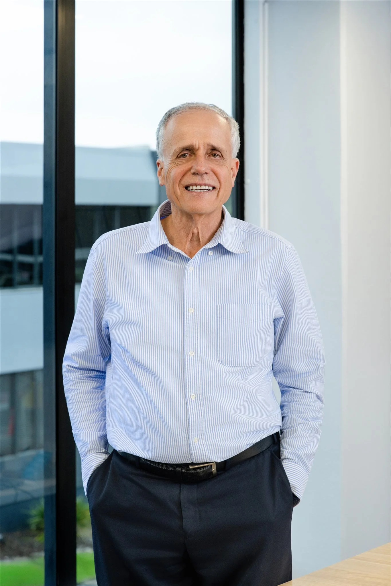 A middle-aged man with gray hair wearing a light blue striped dress shirt and dark pants, standing indoors near large windows, smiling.