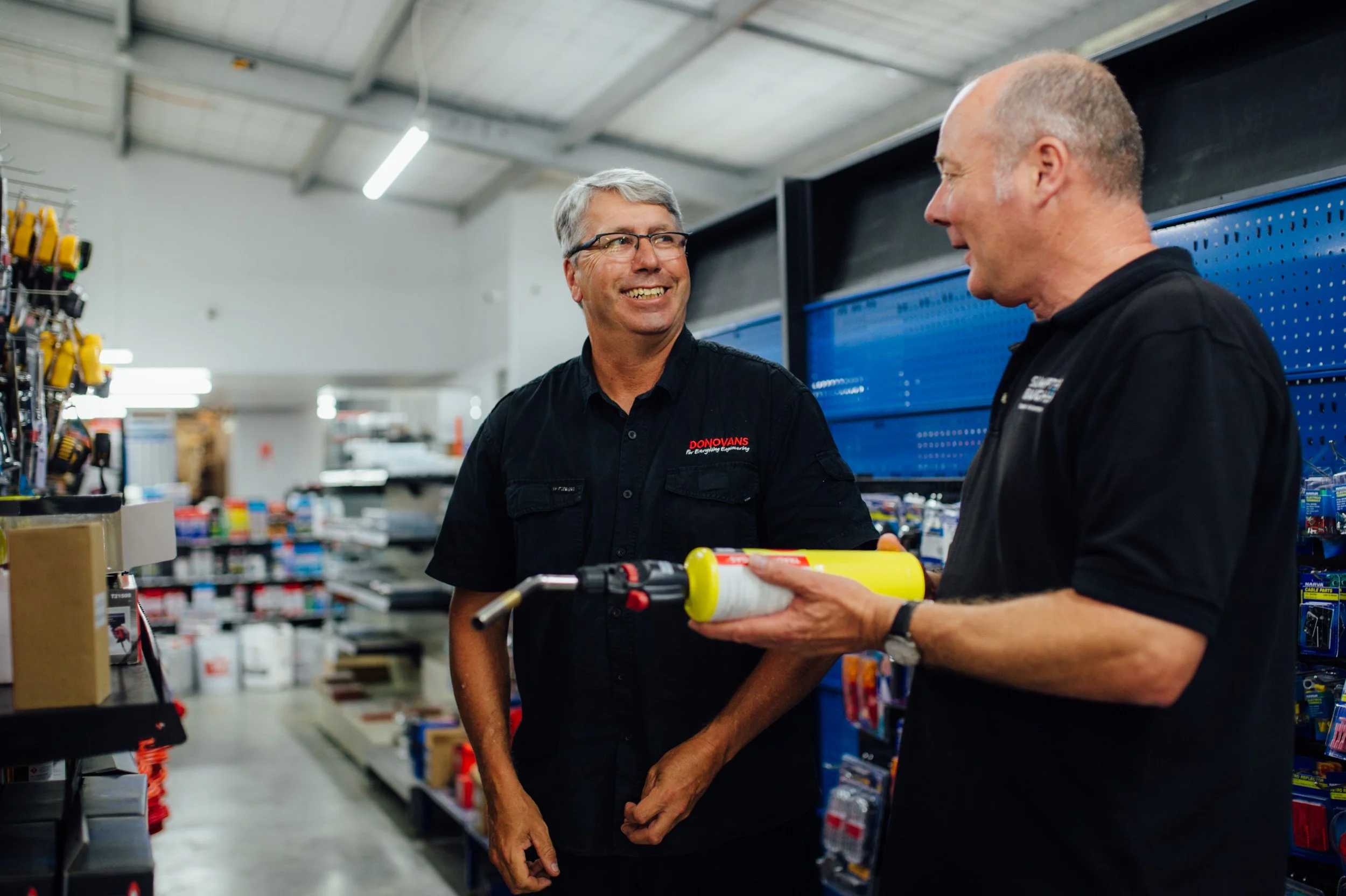 Two men in black work shirts talking in hardware store aisle, one holding a caulking gun.