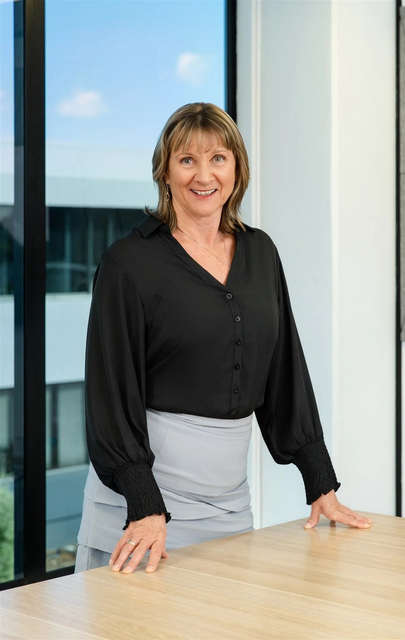 A middle-aged woman with shoulder-length hair standing behind a wooden table in a modern office with large windows and a blue sky outside.