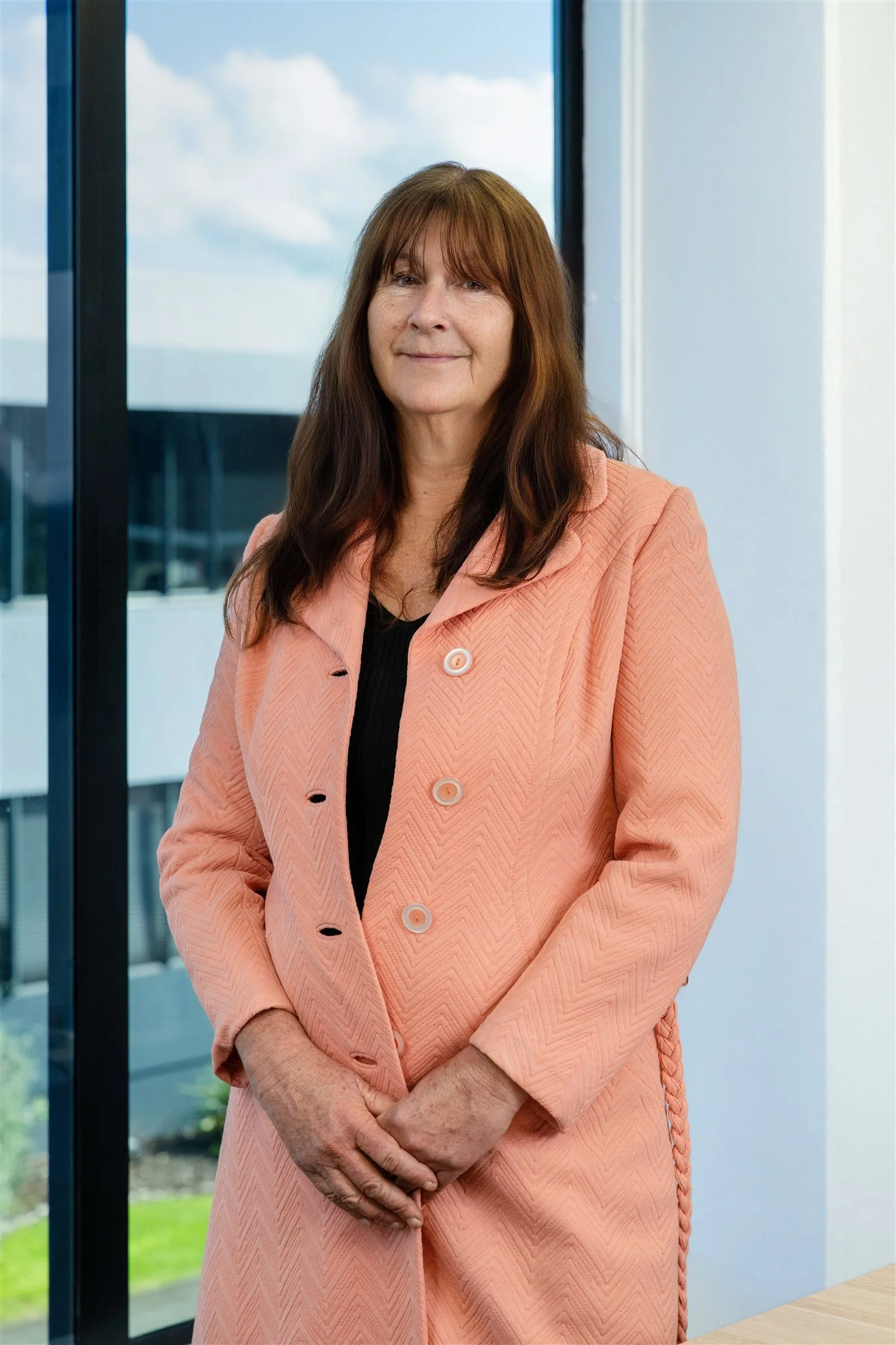 A middle-aged woman with long brown hair wearing a peach-colored blazer standing indoors near large windows with a view of buildings and a partly cloudy sky.