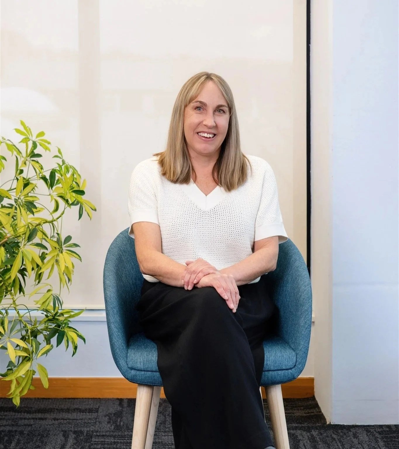 Woman sitting in a blue chair, smiling, in an office setting with a plant to her left.