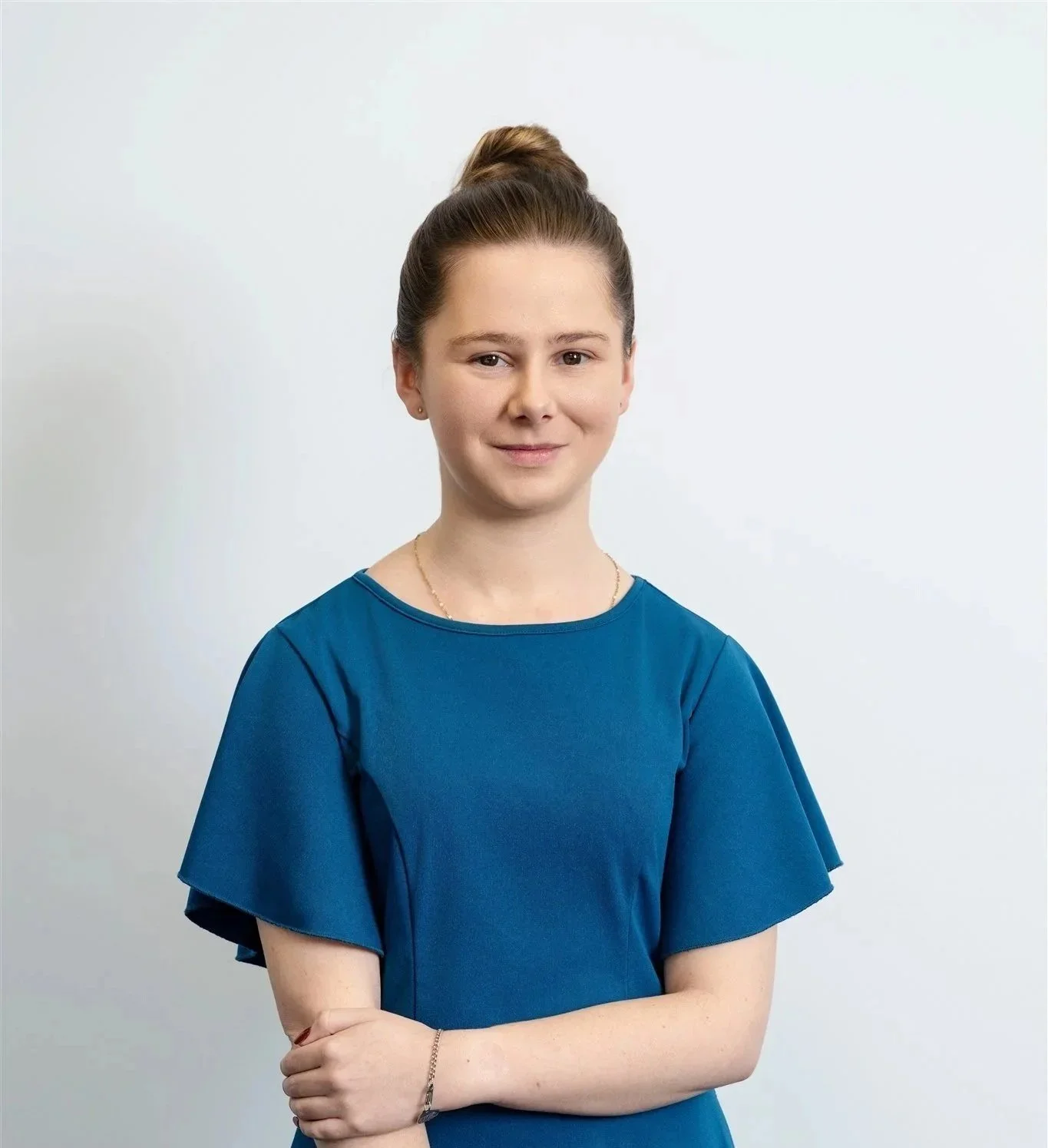 A young woman with brown hair tied in a bun, wearing a blue dress, standing against a plain white background, smiling slightly.